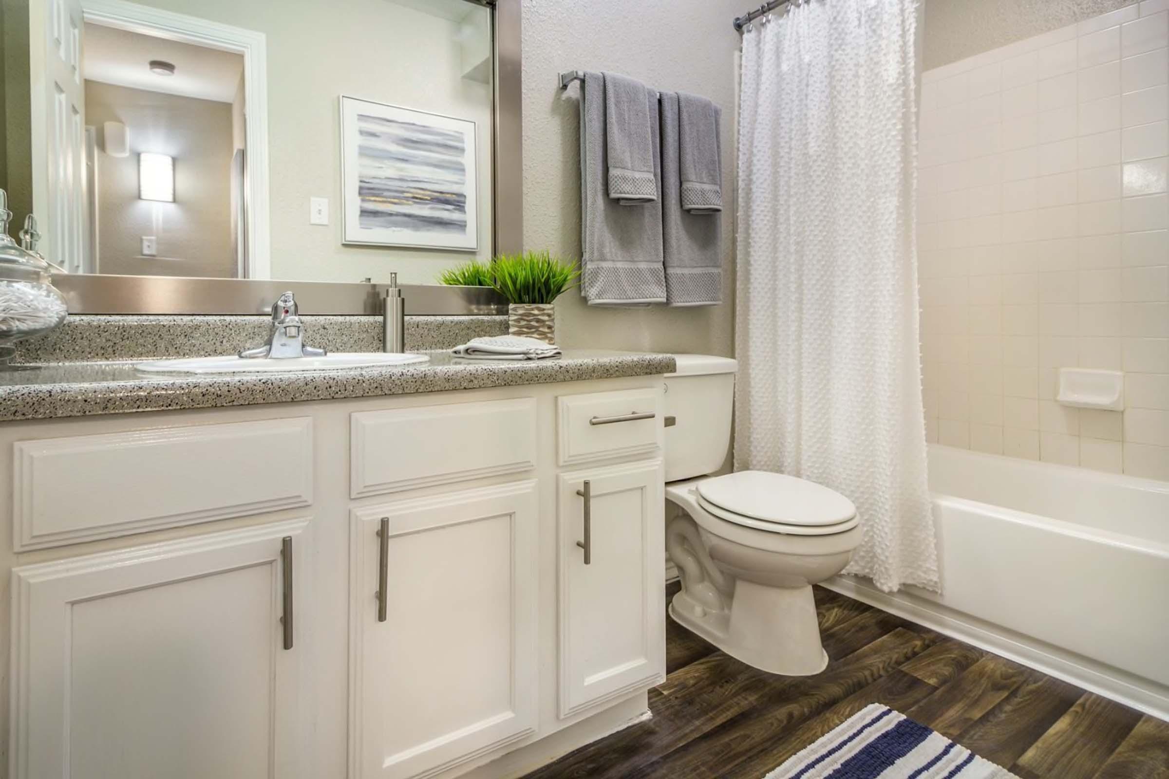Modern bathroom featuring a white vanity with a granite countertop, a mirror, and neatly arranged towels. There is a bathtub with a glass shower curtain, and a small plant accent. The floor is laminate wood, and the walls are a light neutral color, creating a clean and inviting atmosphere.
