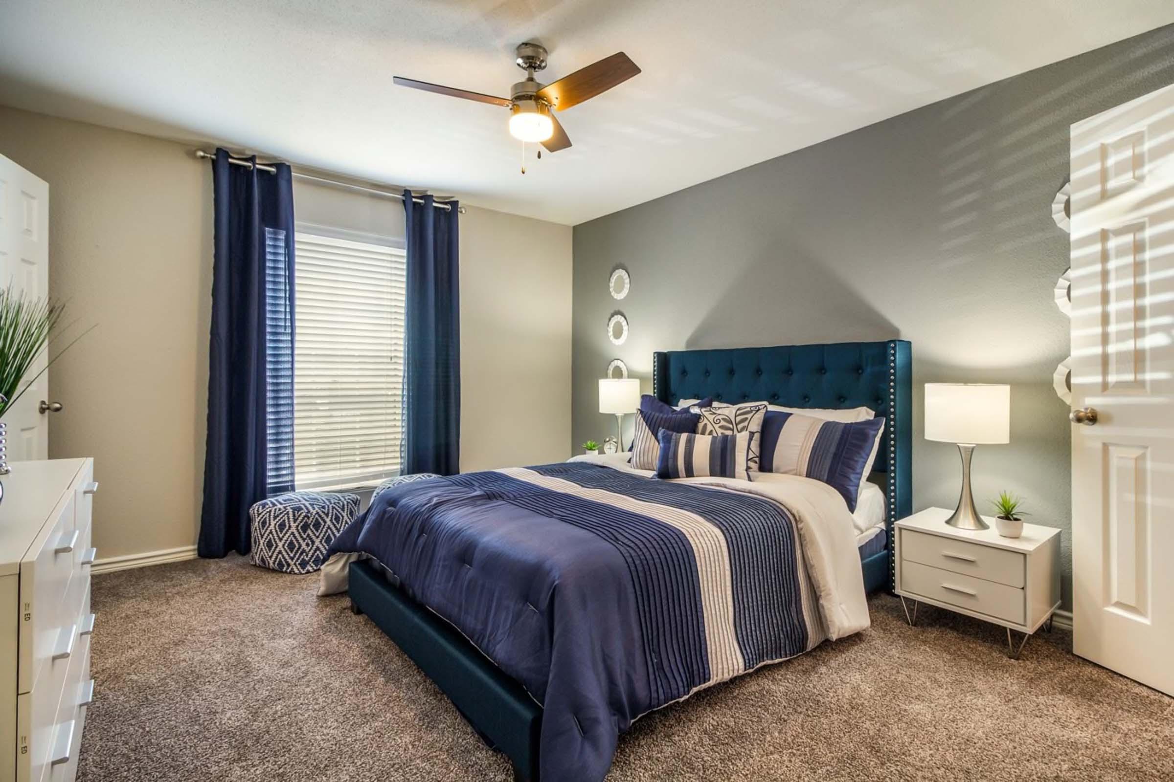 A cozy bedroom featuring a blue tufted bed with decorative pillows and a white throw blanket. The room has a large window with light-filtering curtains, a ceiling fan, and a white dresser. Two bedside lamps illuminate the space, which includes a small decorative plant and a comfortable seating area.