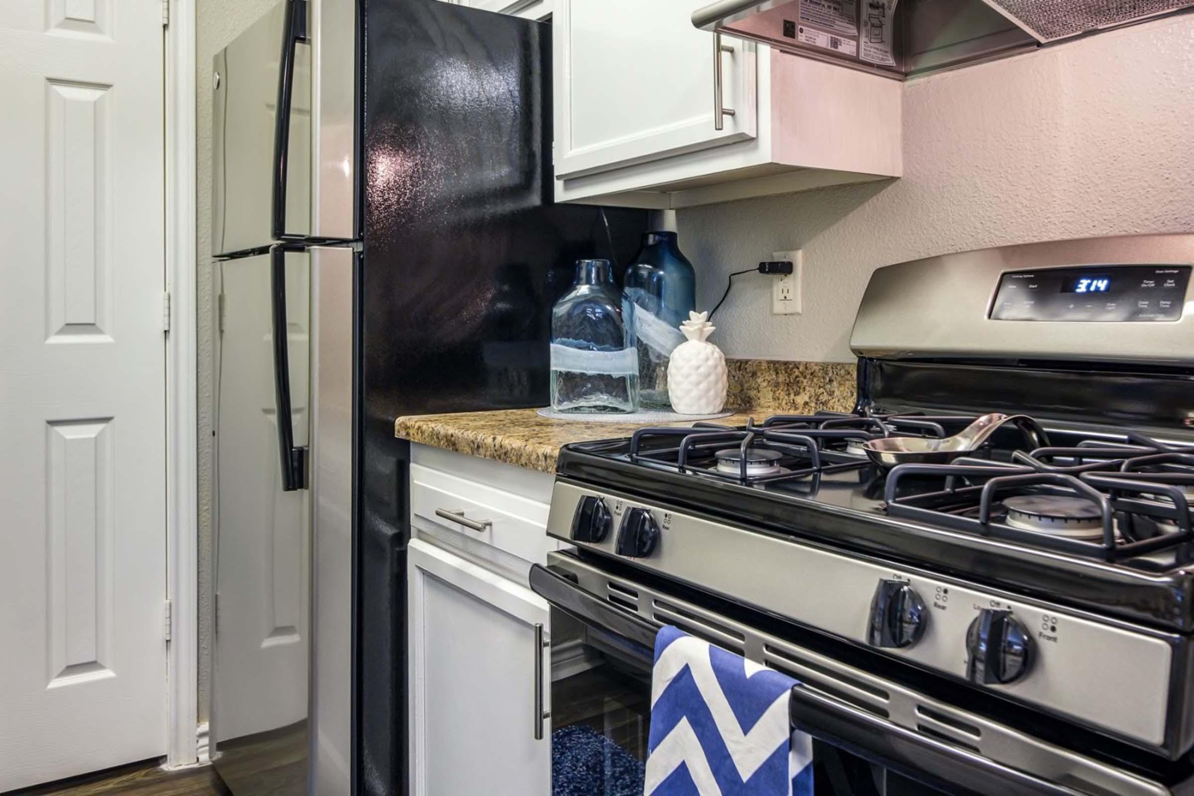 A modern kitchen featuring a black refrigerator, white cabinets, and a stainless steel gas stove. The countertop has a decorative arrangement including glass jars and a pineapple. A blue chevron-patterned towel hangs from the stove, and a door is visible in the background.
