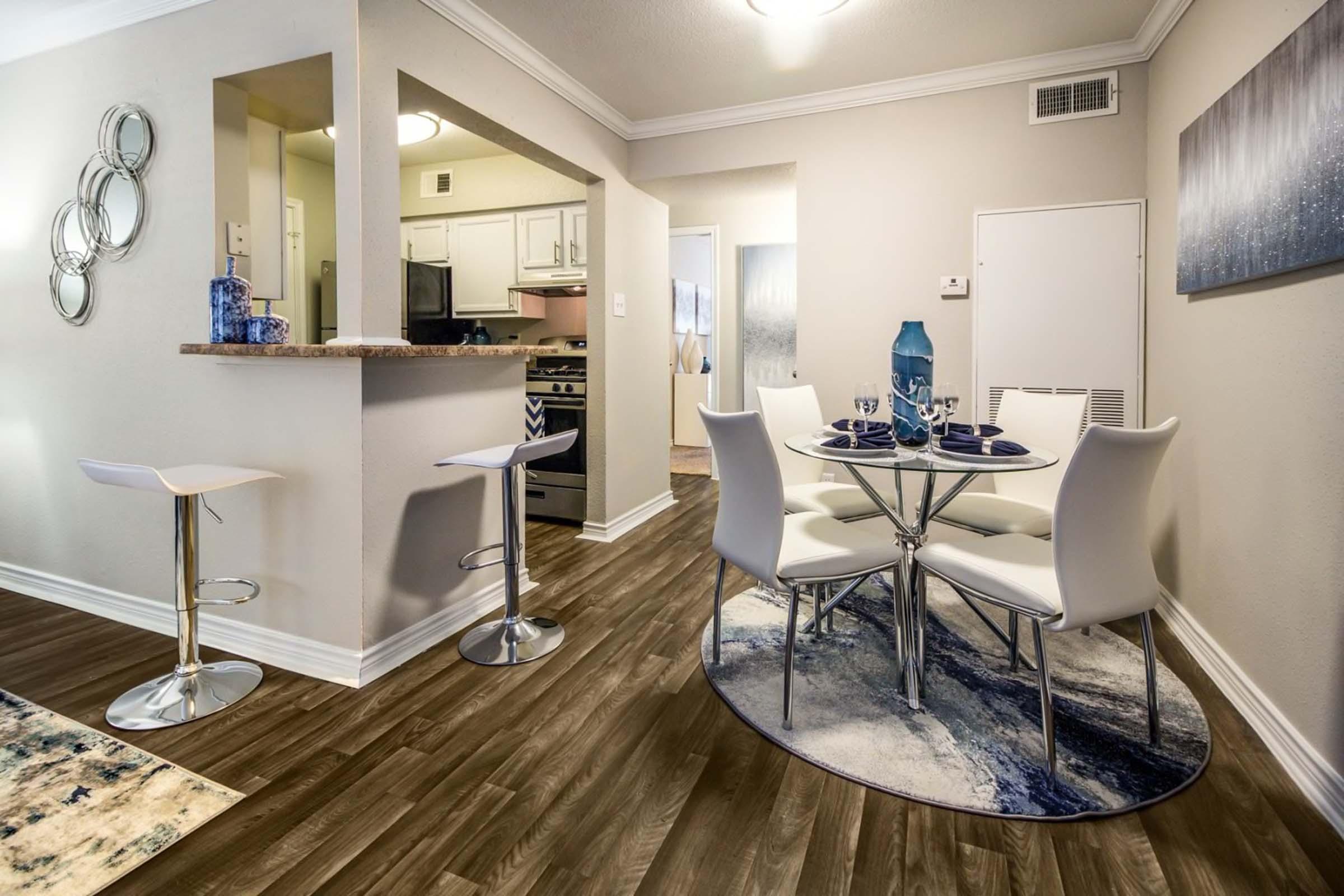 Modern dining area featuring a round glass table with white chairs, set with dishes and glasses. Adjacent kitchen area visible with white cabinets and a bar counter. The floor has a wooden finish, and there are decorative elements like a rug and wall art in soothing colors.