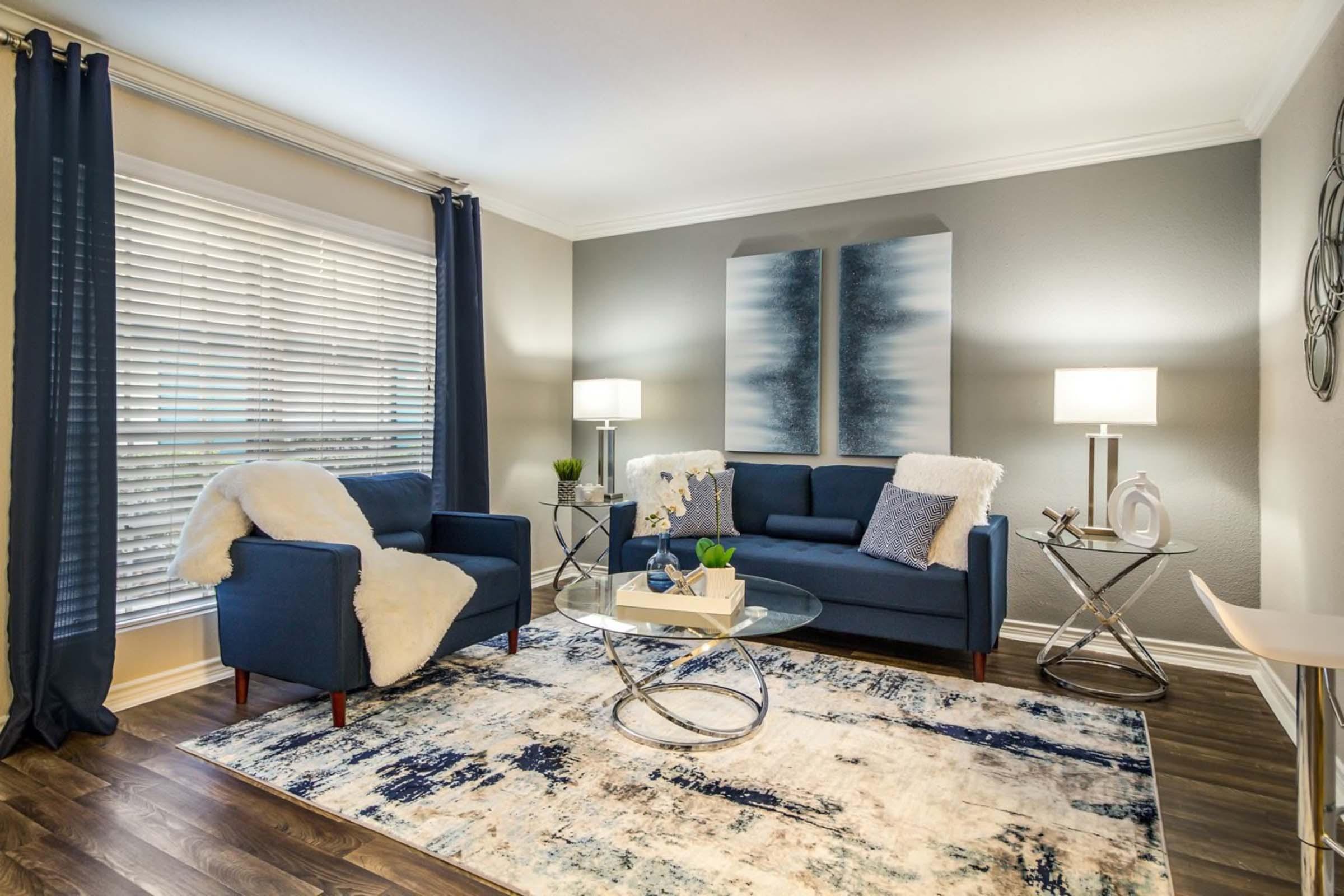 A cozy living room featuring a blue sofa and armchair with white throw blankets, a glass coffee table, and wooden flooring. Soft lighting from two lamps illuminates the room. Modern decor includes an abstract wall art piece and a patterned area rug. Large blinds cover a window, allowing soft natural light.