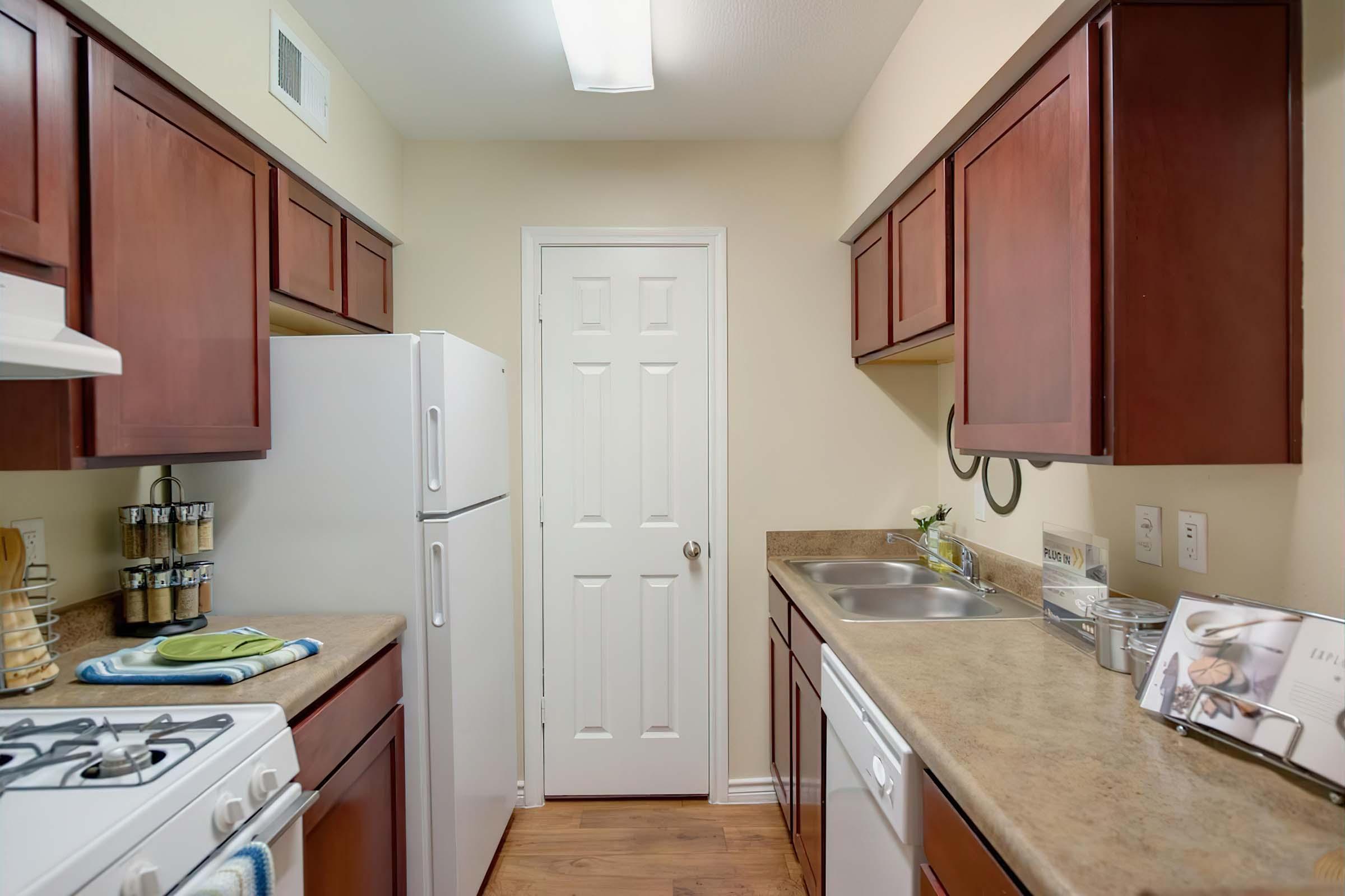 A modern kitchen featuring wooden cabinets, a refrigerator, and a gas stove. The counter is equipped with a sink, decorative items, and kitchen utensils. A closed door can be seen at the end of the kitchen, and the overall layout is bright and inviting, showcasing a clean and organized space.