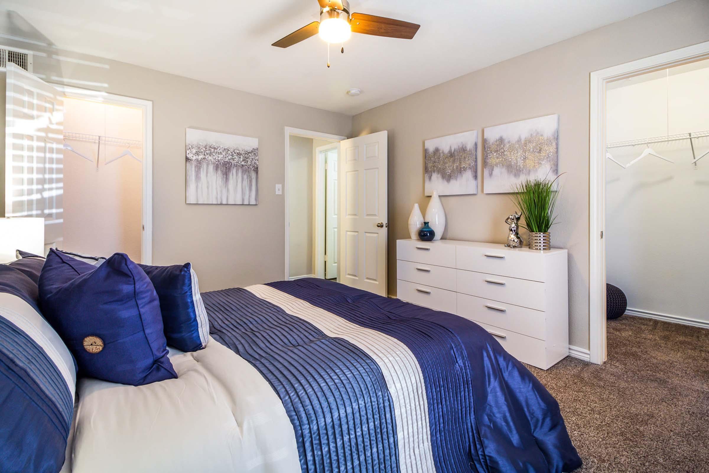 A cozy bedroom featuring a neatly made bed with blue and white bedding. The room has light beige walls, a ceiling fan, and framed artwork on the wall. A white dresser is positioned against one wall, and a door leads to a closet, adding to the spacious feel of the room.