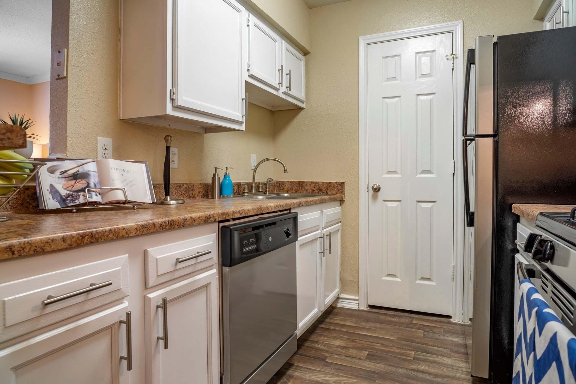 A modern kitchen featuring white cabinets, a granite countertop, and stainless steel appliances. The sink is under the window, with a dishwasher next to it. A black refrigerator is visible, along with an oven. The floor has a wooden appearance, and the walls are painted a light beige color.