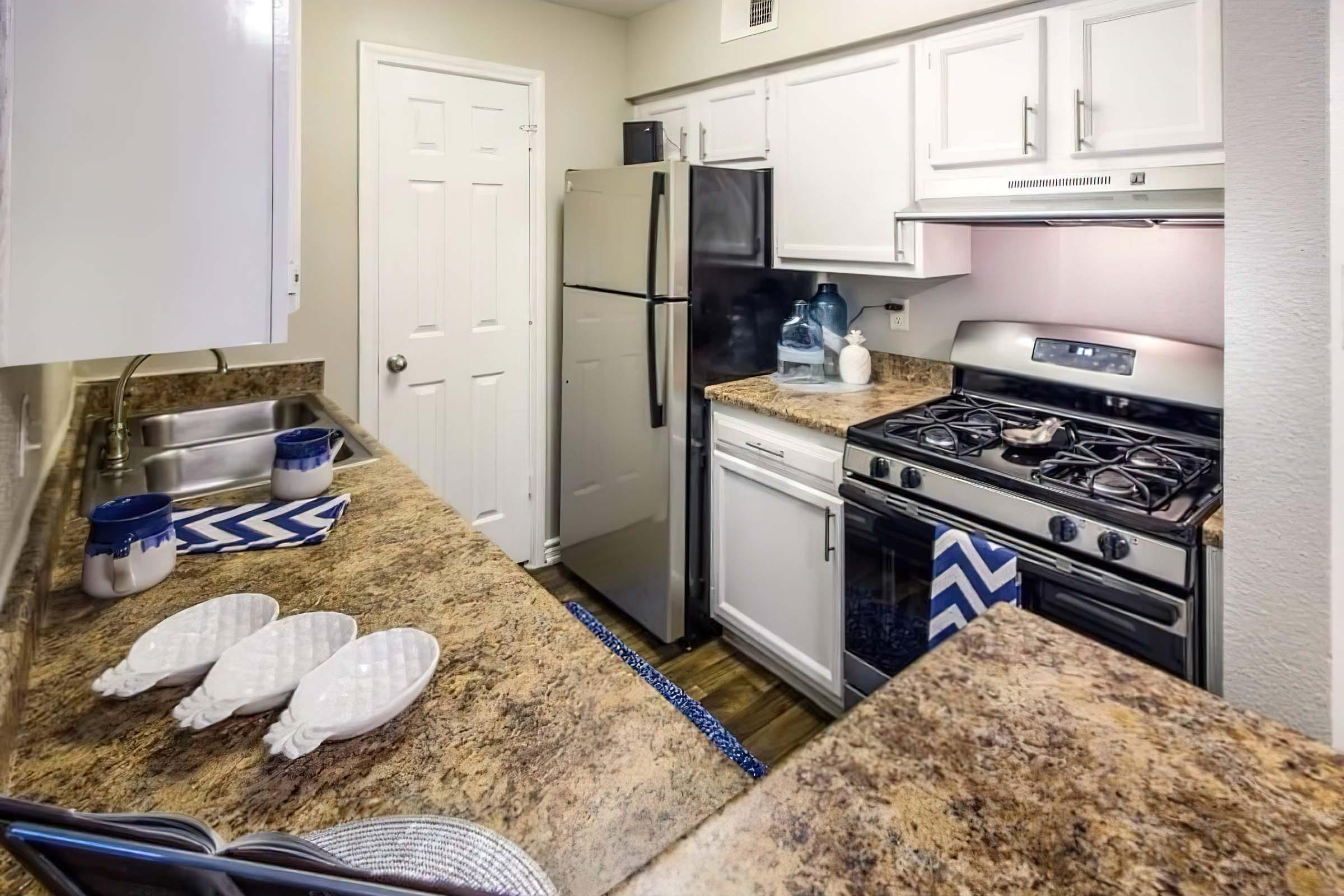 A modern kitchen featuring light-colored cabinets, a black refrigerator, a gas stove with an oven, and a stainless steel sink. The countertops are brown with a textured finish. Decorative plates and blue-striped dish towels are on the counter. A door leads to another space, and there are water bottles on the counter.