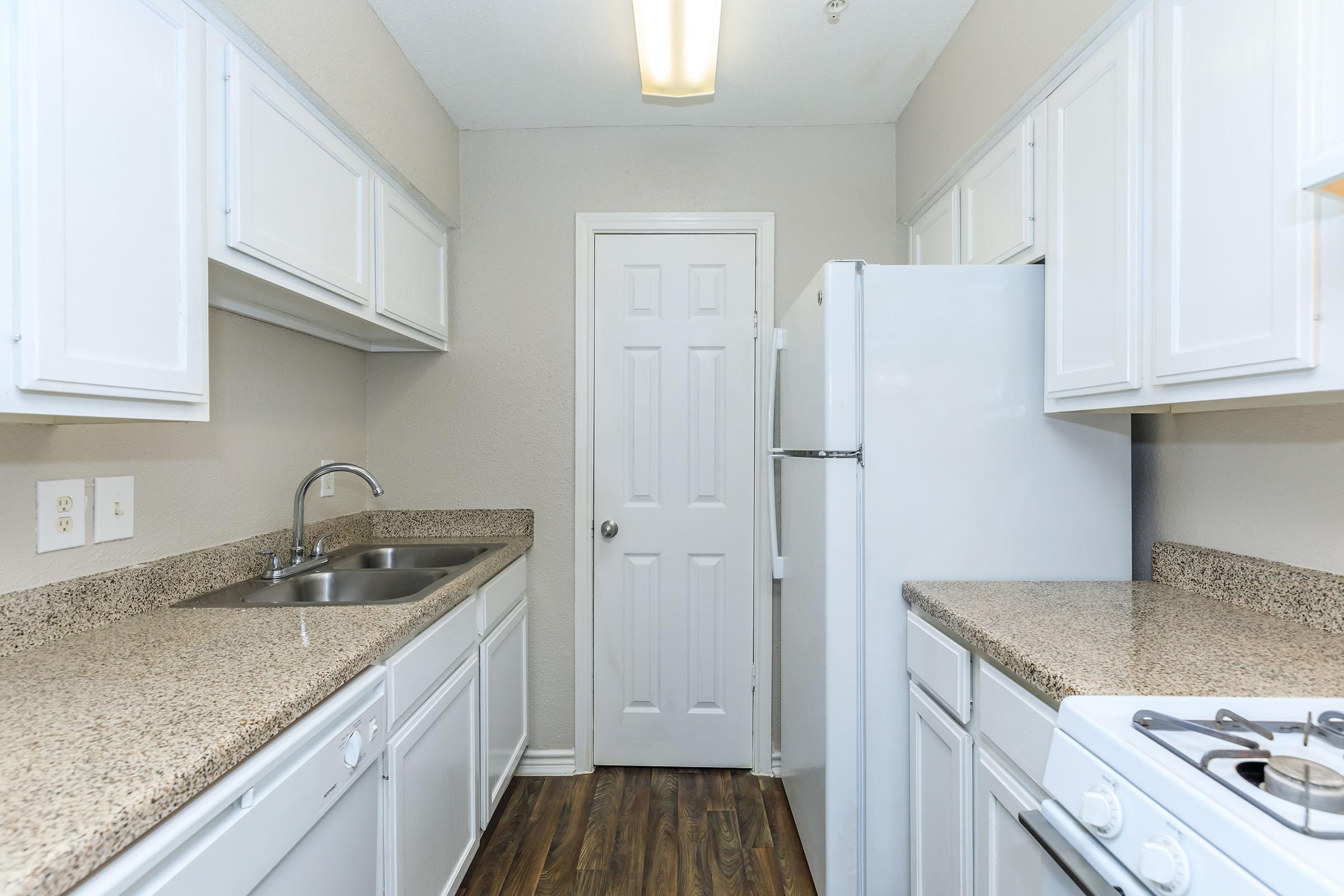 A modern kitchen featuring white cabinets, a granite countertop, a stainless steel sink, and a refrigerator. The space includes a door leading to another area and a gas stove on the right. The walls are painted in a neutral color, and the flooring resembles wood for a warm ambiance.