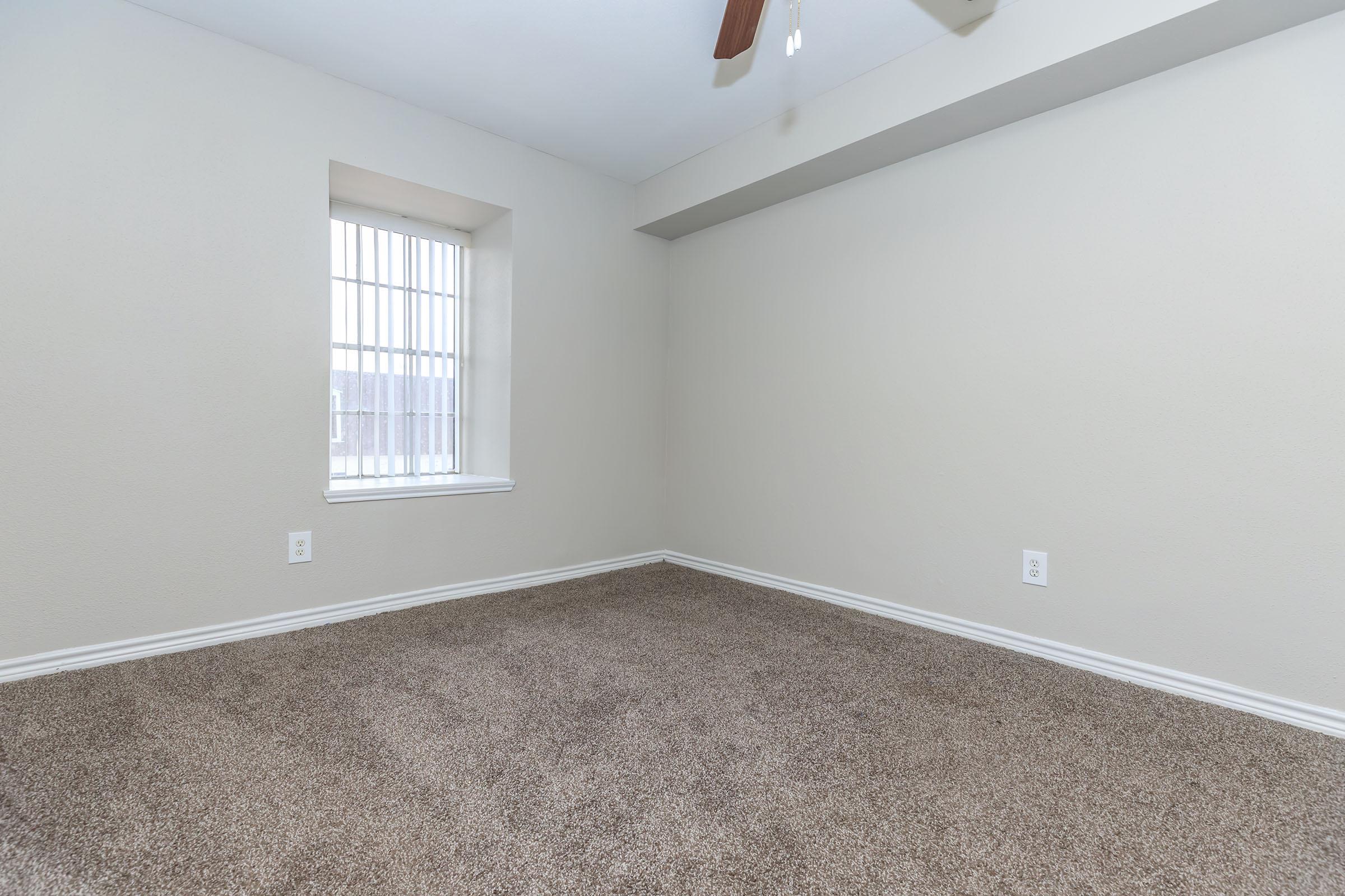 Empty room with beige walls and light brown carpet. There's a small window with white blinds on one side, and a ceiling fan above. The room has a spacious feel with no furniture or decorations, showcasing the clean and simple design.