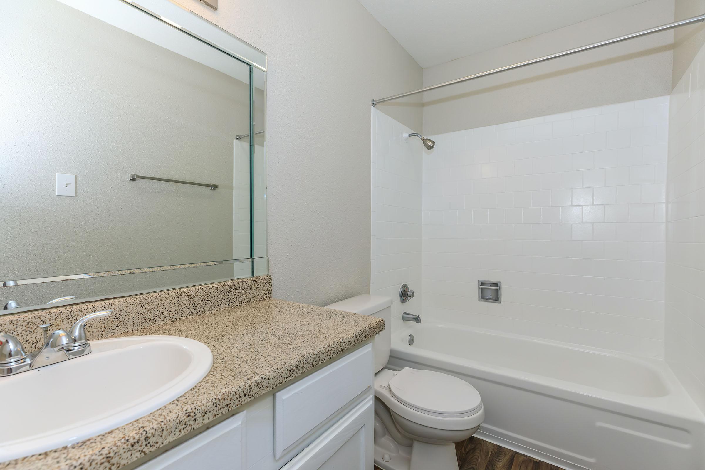 A clean and modern bathroom featuring a white bathtub with a chrome shower rod, a granite countertop with a white sink, and a large mirror above the sink. The walls are painted in a light color, and the floor has a wood-like appearance, creating a bright and spacious atmosphere.