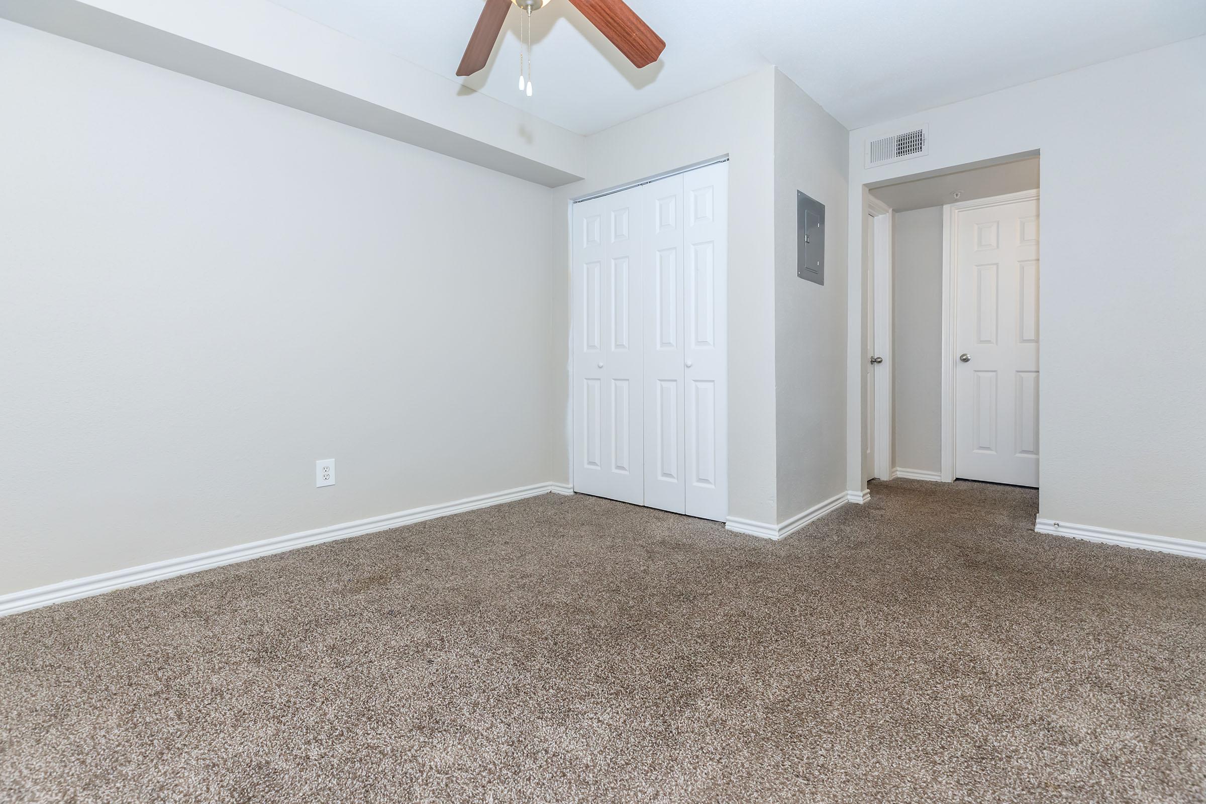 A spacious and empty room with light gray walls and plush brown carpet. There is a ceiling fan with wooden blades, a double-door closet on one side, and a doorway leading to another room. The overall ambiance is bright and neutral, suitable for various interior design styles.