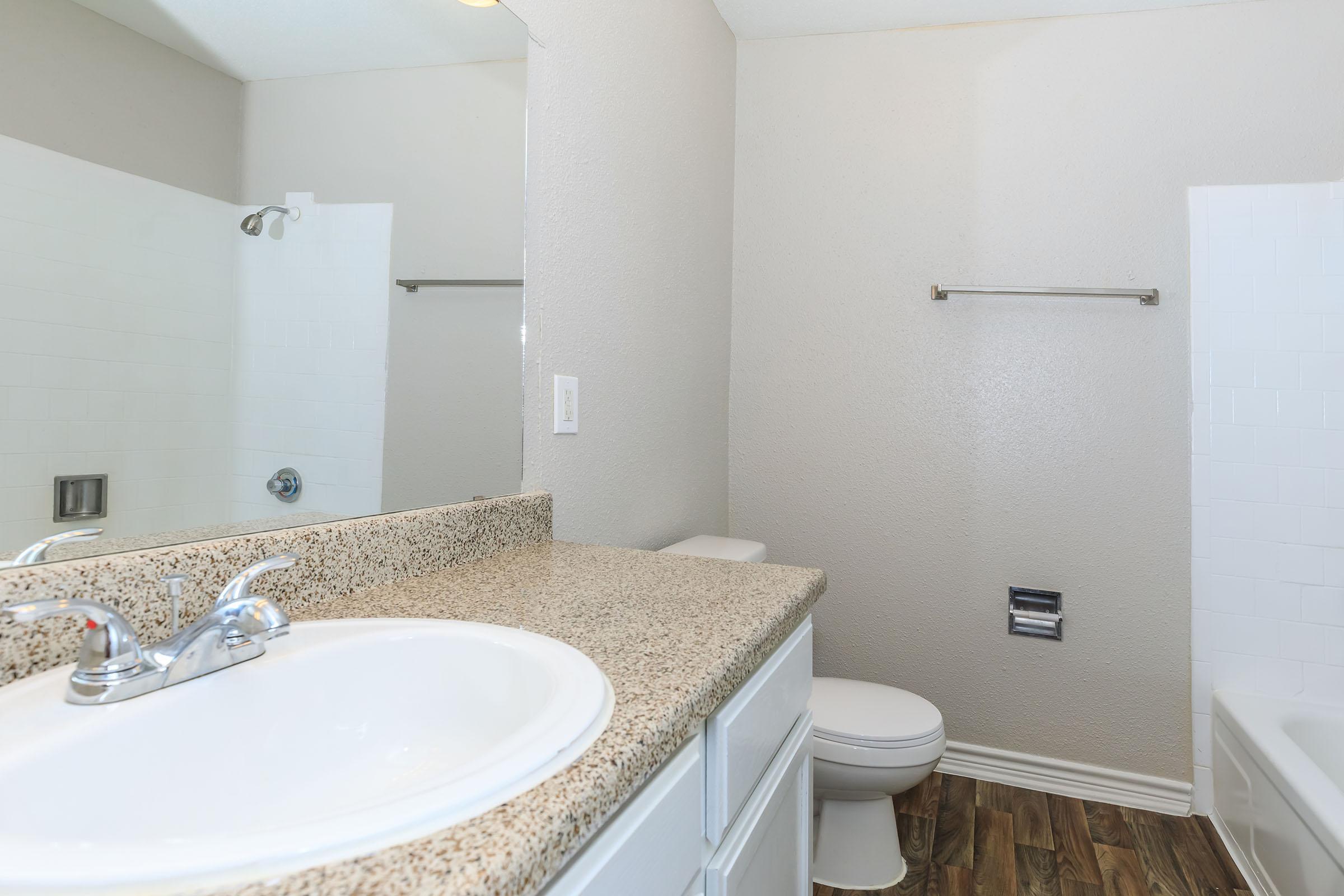 A clean and modern bathroom featuring a white sink with a granite countertop, a toilet, and a simple shower area. Light-colored walls and wood-like flooring enhance the spacious feel. A mirror above the sink reflects the room, adding to the bright and airy atmosphere.