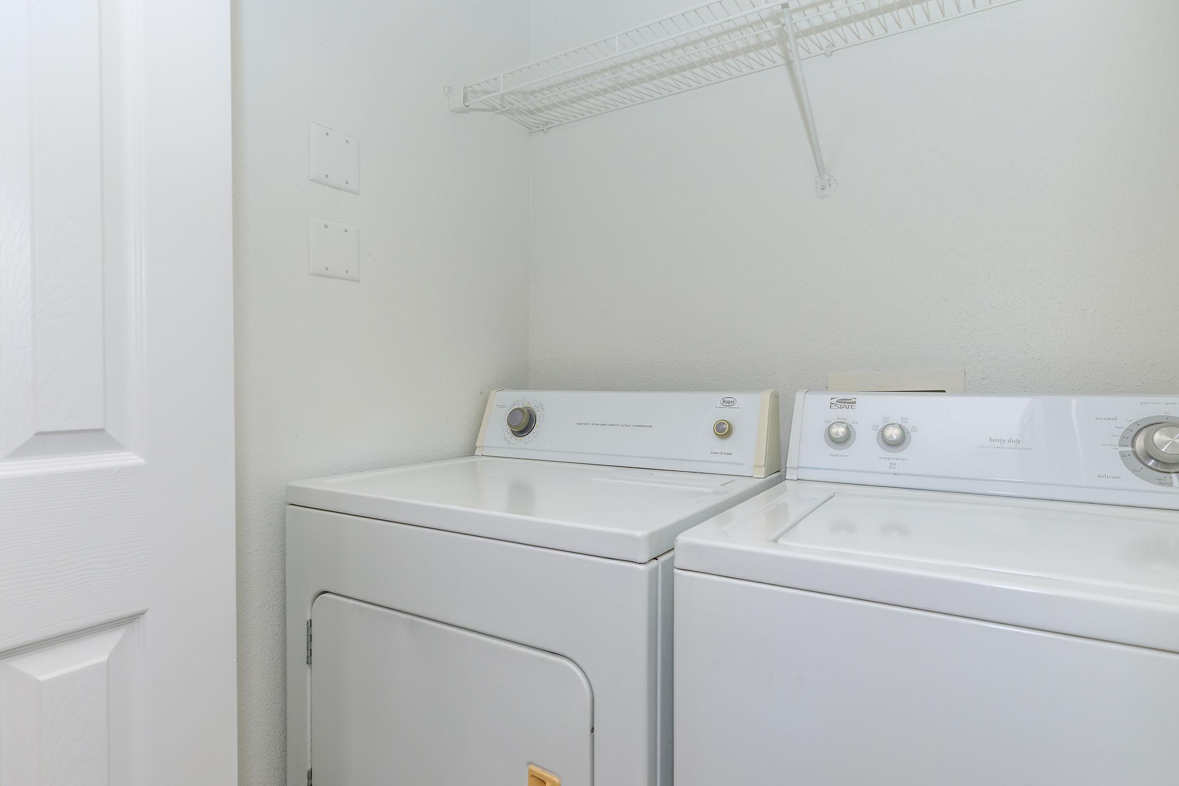 A laundry room featuring a stacked white washer and dryer on the left and right against a plain wall. Above the appliances, a metal wire shelf for storage is mounted. The room has a light and airy feel with a door partially visible on the left.