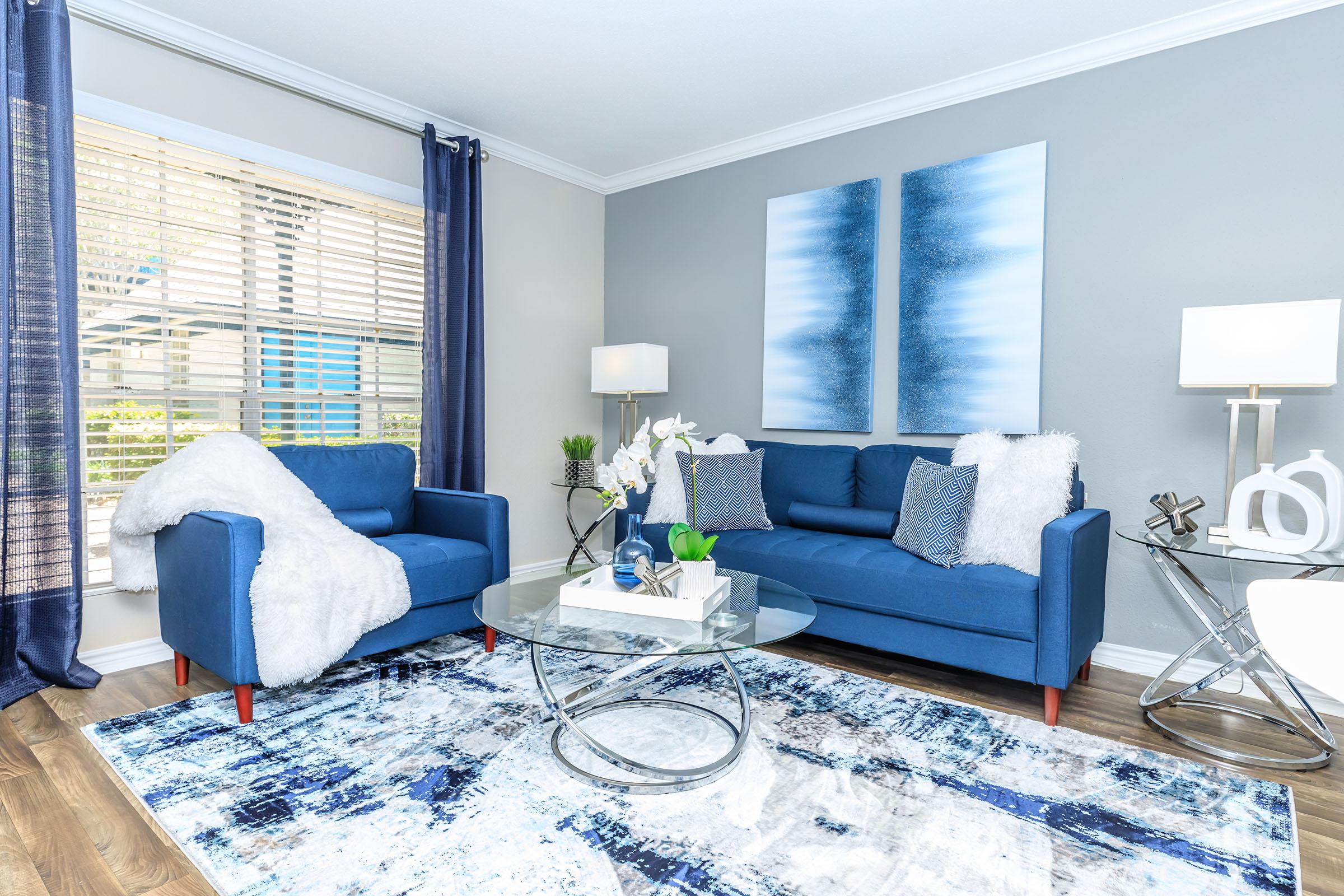 A cozy living room featuring blue furniture, including a sofa and armchair, accented with throw pillows and a soft white blanket. A glass coffee table sits on a patterned area rug, while curtains frame a window with natural light filtering in. Abstract blue artwork hangs on the wall, enhancing the modern aesthetic.