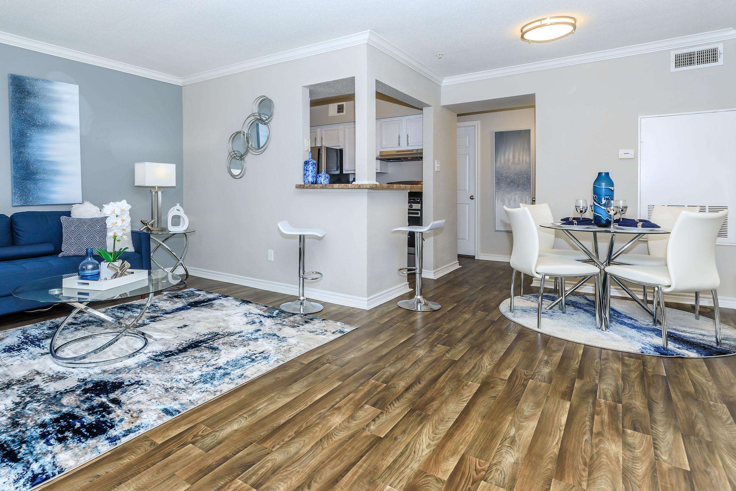 A modern living space featuring a blue couch, round dining table with white chairs, and a light wood floor. Decor elements include a rug with blue and neutral tones, a wall mirror, and decorative art. A kitchen area is visible in the background, showcasing cabinets and appliances.