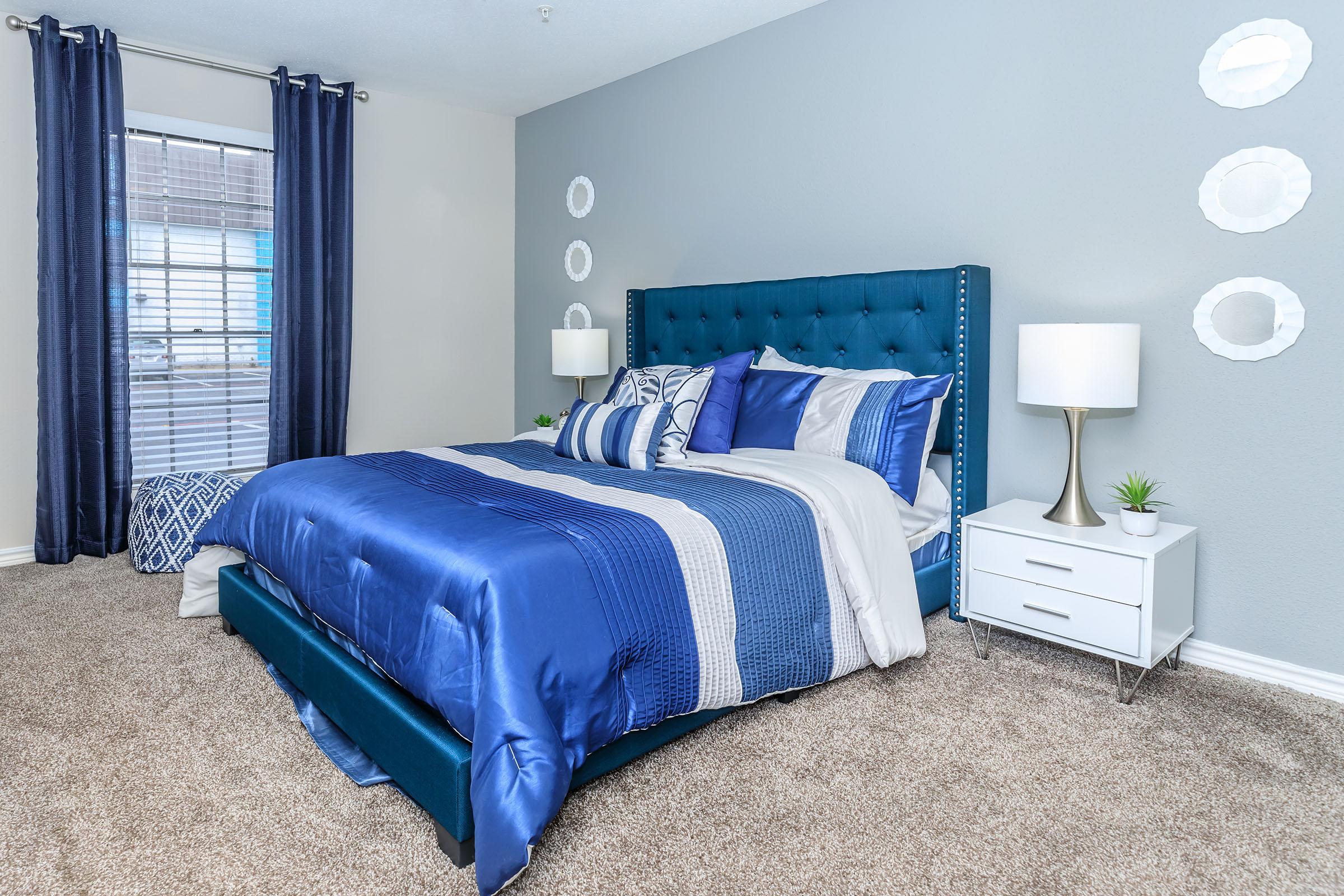 A cozy bedroom featuring a teal upholstered bed with blue and white bedding, two decorative pillows, and matching curtains. A bedside table with a lamp and a small plant sits next to the bed. The room has light-colored walls, plush carpet, and circular wall decor, creating a welcoming and stylish atmosphere.