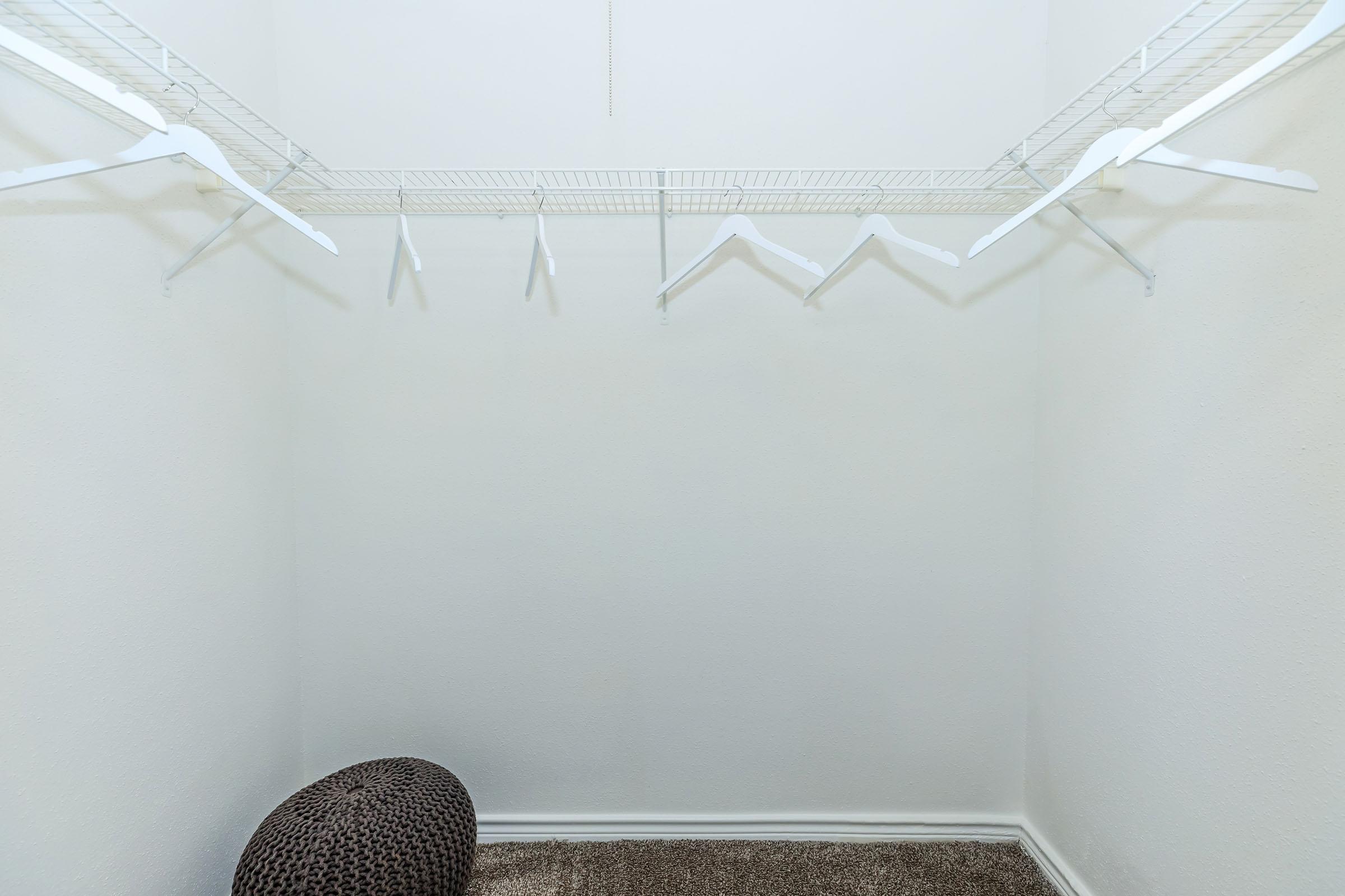 An empty closet with a white wire shelving unit on the wall. Several white hangers are hanging from the shelf, and there is a round, textured brown pouf on the carpeted floor. The walls are plain and painted white, giving the space a clean, minimalist look.