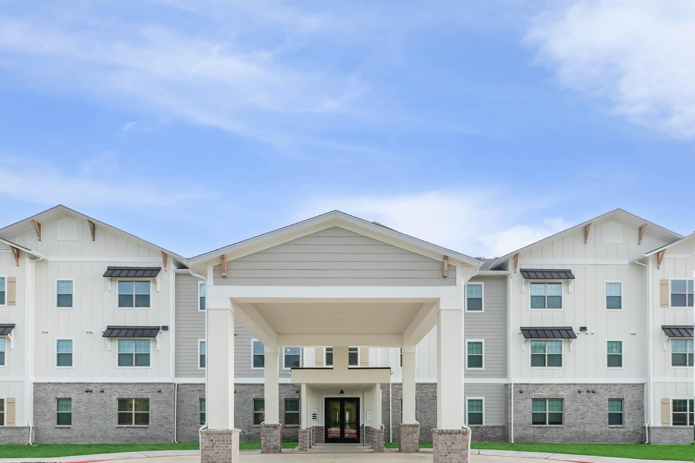 A modern multi-story building with a light-colored exterior and stone accents. The entrance features a large covered area supported by white pillars, with windows visible on the upper floors. The sky above is clear and blue, suggesting a pleasant day. Green grass surrounds the entrance area.