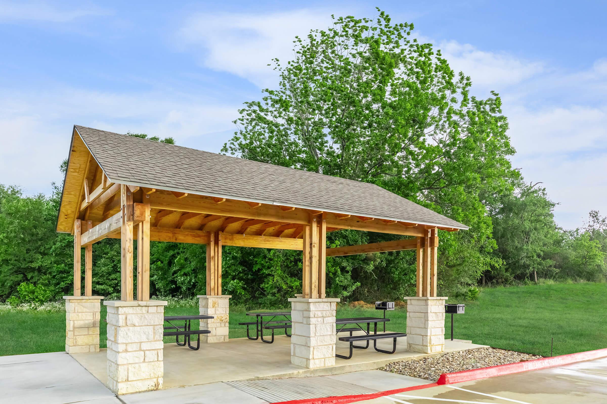 A wooden picnic shelter with a slanted roof, supported by stone columns, located on a paved area. There are several picnic tables underneath, and it is set against a backdrop of green trees and a clear blue sky. The area appears inviting for outdoor gatherings and activities.