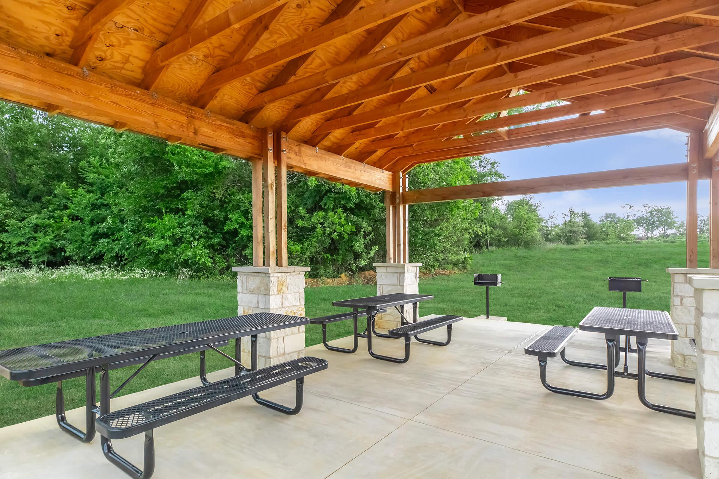 An outdoor picnic shelter featuring wooden beams and stone columns, with several black metal picnic tables arranged beneath it. The surrounding area consists of green grass and trees, creating a tranquil setting for gatherings or meals.