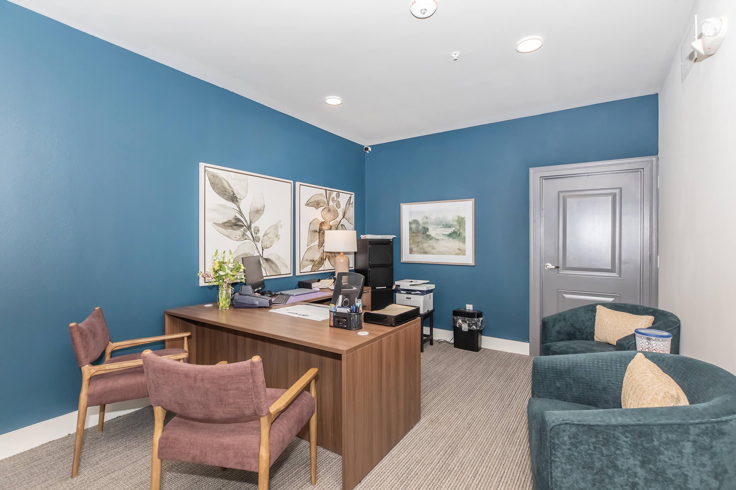 A small office space featuring a wooden desk with paperwork and a lamp, a printer, a wastebasket, and two comfortable seating areas with chairs. The walls are painted blue and adorned with framed artwork. The floor is carpeted, creating a welcoming and professional atmosphere.