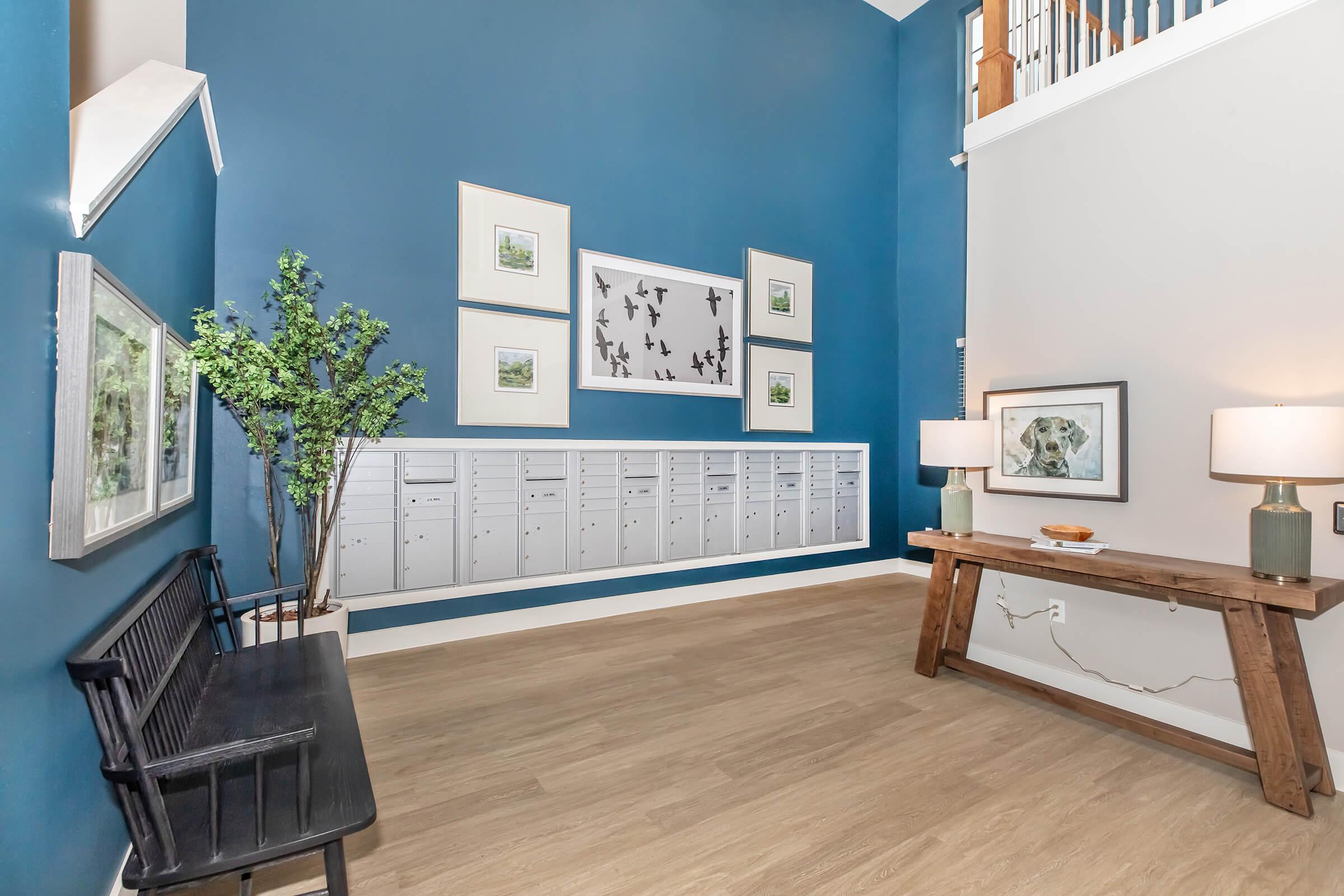 A modern entryway featuring a blue accent wall with framed artwork of birds. There’s a row of mailboxes mounted on the wall, a wooden bench on the left, and a rustic wooden table with a decorative item on top. Two lamps on the table provide soft lighting, enhancing the welcoming atmosphere.