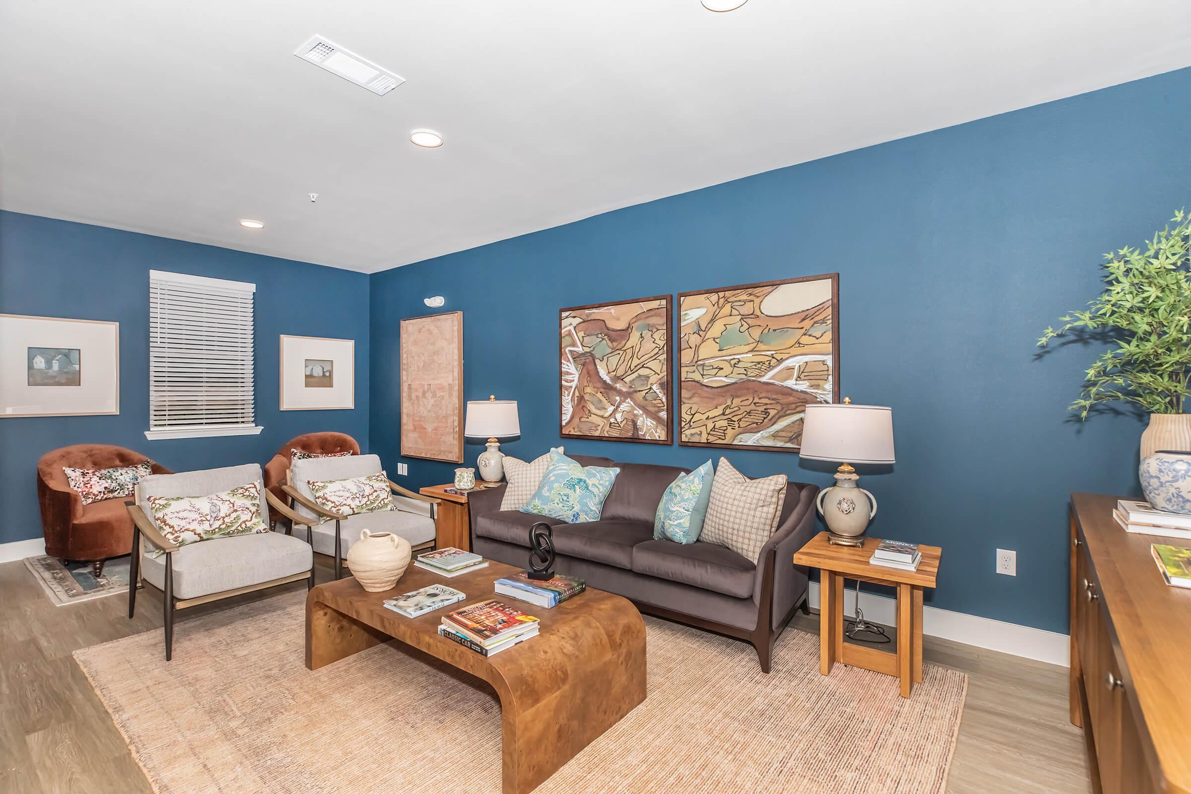 A cozy living room with a dark blue accent wall. The space features a brown sofa with patterned pillows, two stylish armchairs, and a wooden coffee table topped with books. There are decorative lamps, artwork on the walls, and a plant in the corner, creating a warm and inviting atmosphere.