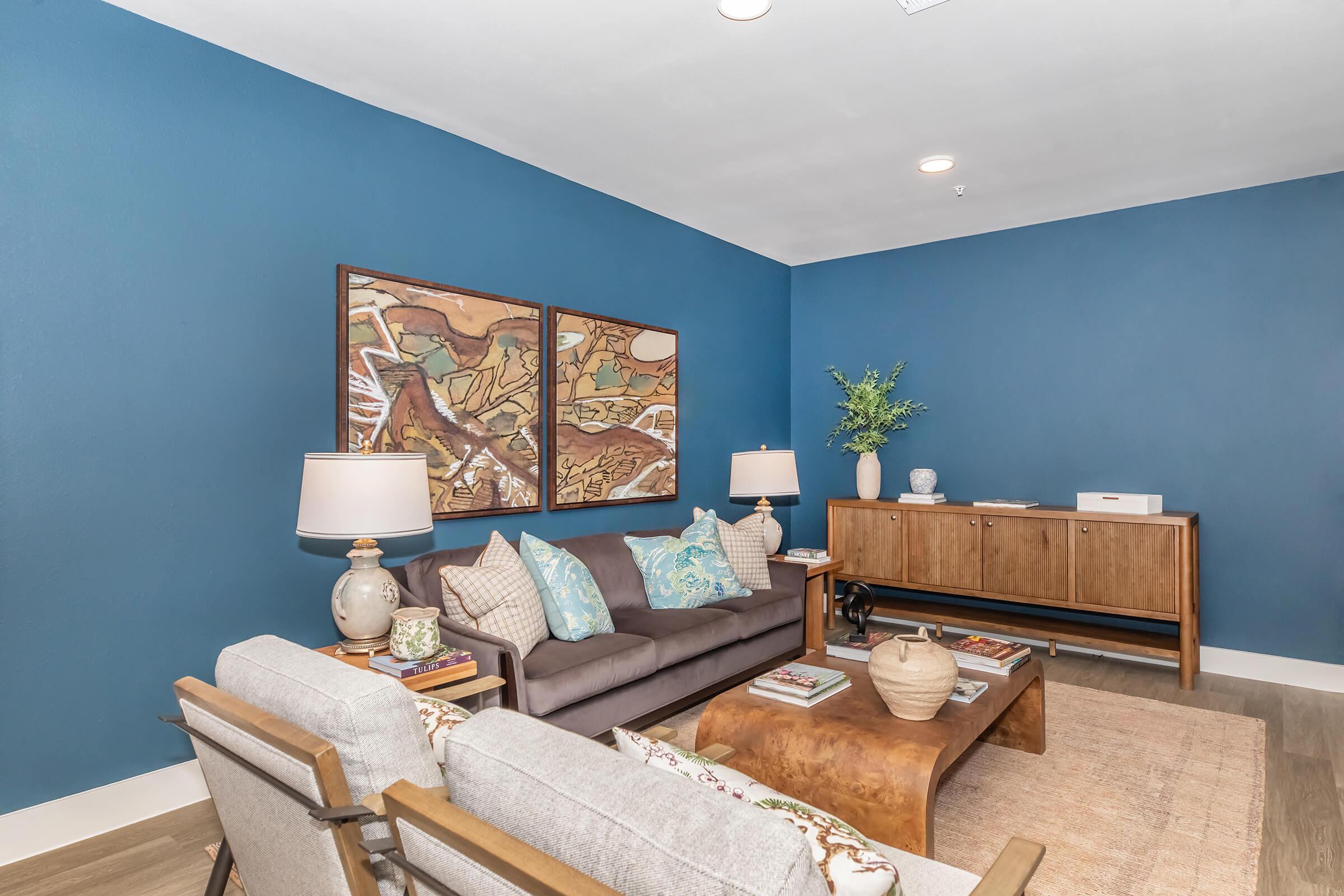 A cozy living room featuring a dark blue accent wall, a stylish gray sofa with decorative pillows, and a pair of modern lamps. Two framed art pieces hang above a wooden sideboard, adorned with a vase of greenery and various decorative items. Natural light enhances the inviting atmosphere.