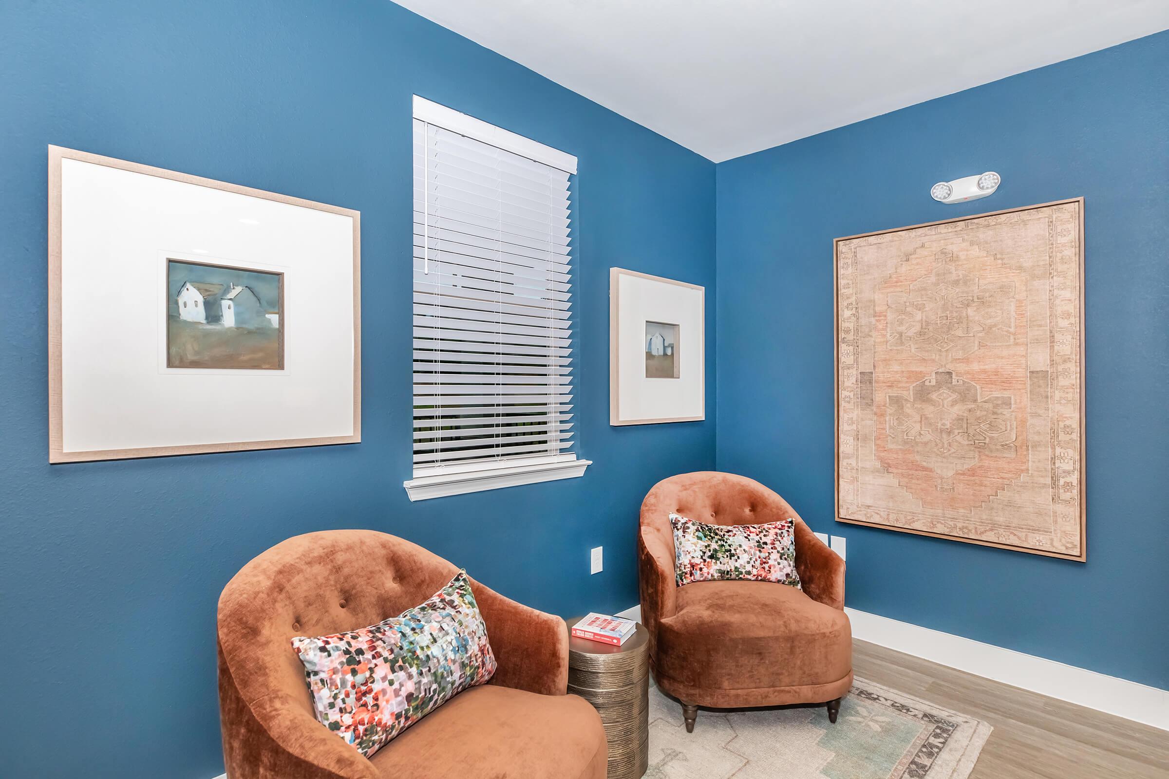 A cozy corner of a room featuring two brown upholstered chairs with floral pillows, framed art on blue walls, and a large map-like artwork. A window with blinds lets in light, and a small table beside the chairs holds a magazine. The space has a warm and inviting atmosphere.