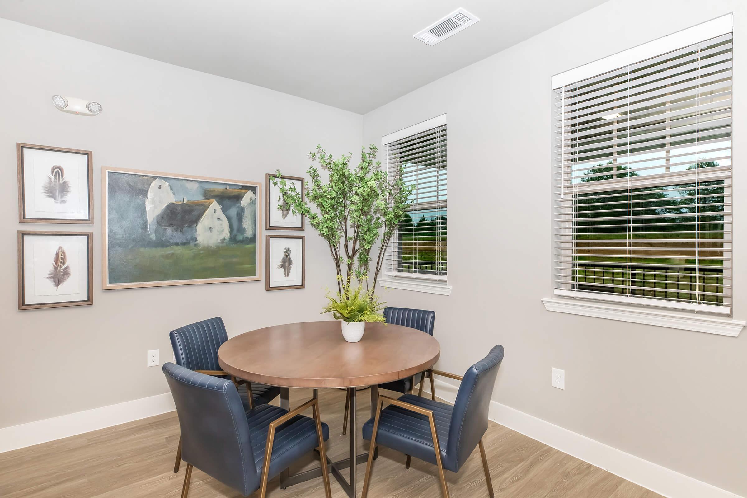 A bright dining area featuring a round wooden table surrounded by four blue chairs. Decor includes framed artwork on the walls, showcasing a landscape painting and feather prints. A green plant in a white pot adds a touch of nature, with large windows letting in natural light.