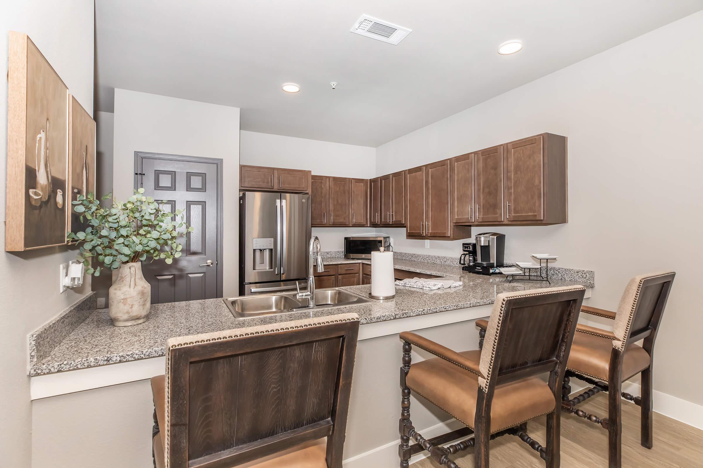 A modern kitchen featuring wooden cabinetry, a stainless steel refrigerator, a microwave, and a granite countertop with bar seating. There are two high-backed chairs at the counter, and a decorative plant in a vase adds a touch of greenery to the space. The walls are painted in a light, neutral color.
