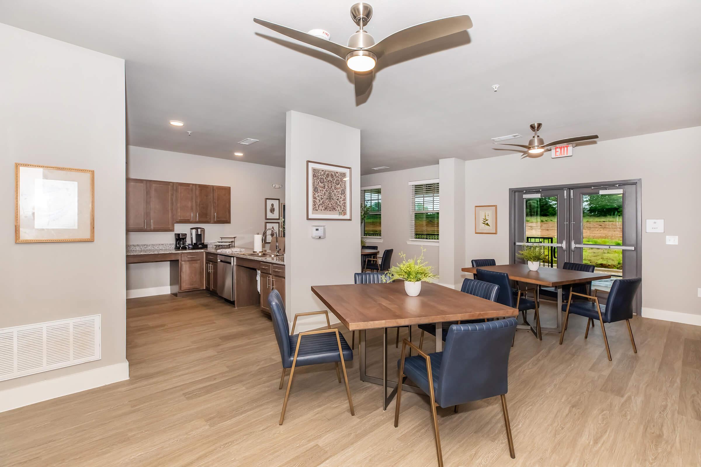 Modern kitchen and dining area featuring a table with dark chairs, a well-equipped kitchen with wooden cabinets, and large windows looking out onto greenery. The space is well-lit with ceiling fans, creating a warm and inviting atmosphere.