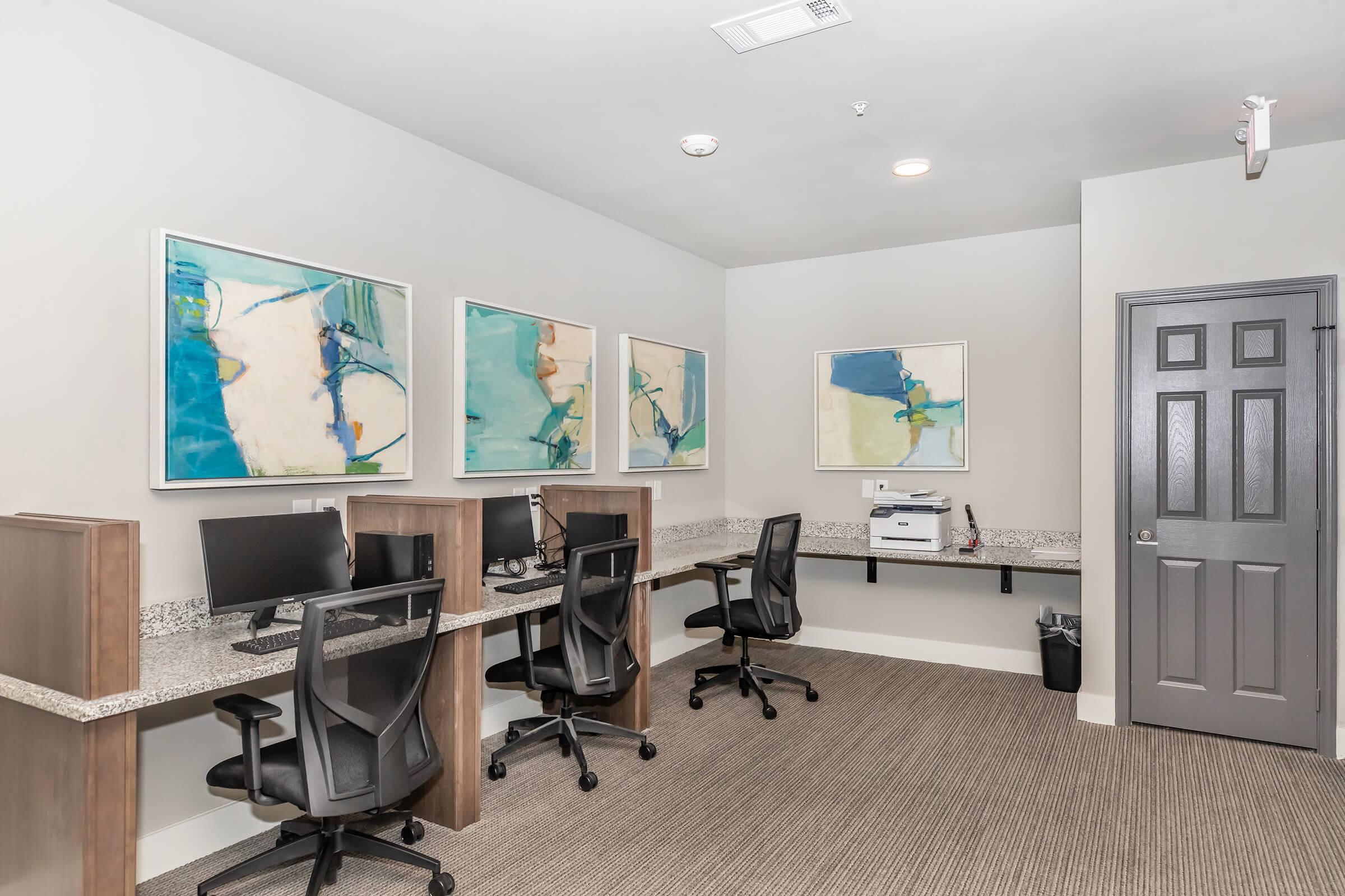 A modern office space featuring two computer desks with black office chairs. There are three abstract art pieces on the wall above the desks, and a printer on the counter. The room has neutral-colored walls and a door on the right. Soft lighting enhances the professional atmosphere.
