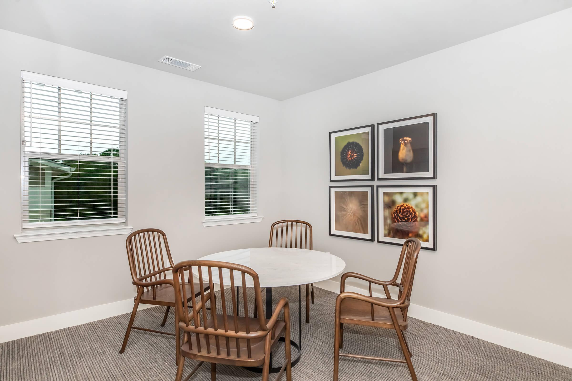 A small meeting room featuring a round marble table surrounded by four wooden chairs. The walls have large framed photographs of nature, including textures and patterns. There are two windows with white blinds letting in natural light, and the room has a neutral color scheme with cozy carpeting.