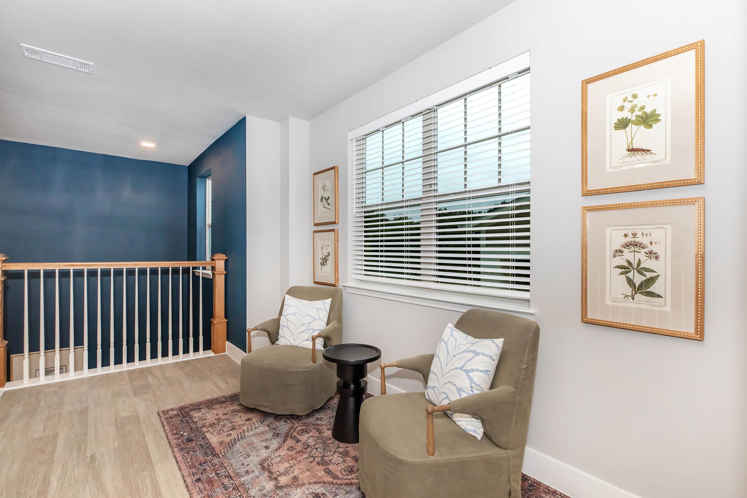 A cozy indoor space featuring two comfortable armchairs with patterned cushions, a small side table between them, and decorative wall art. A white railing borders the area, with a blue accent wall in the background and ample natural light streaming through large windows.