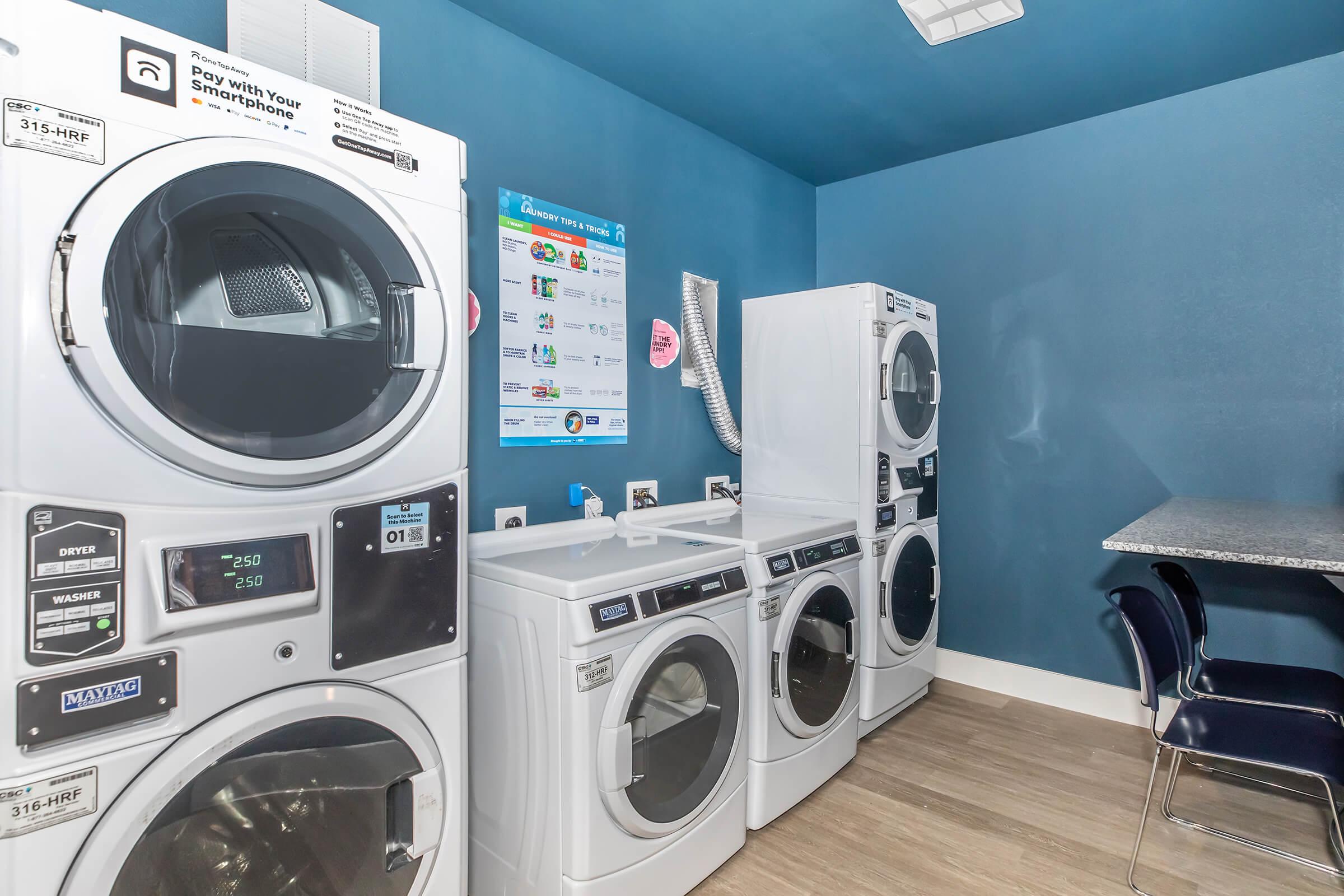 Laundry room with multiple washing machines and dryers lined against a blue wall. A folding table is visible, along with a bulletin board displaying laundry instructions and amenities. The space appears modern and well-lit, designed for convenience and efficiency.