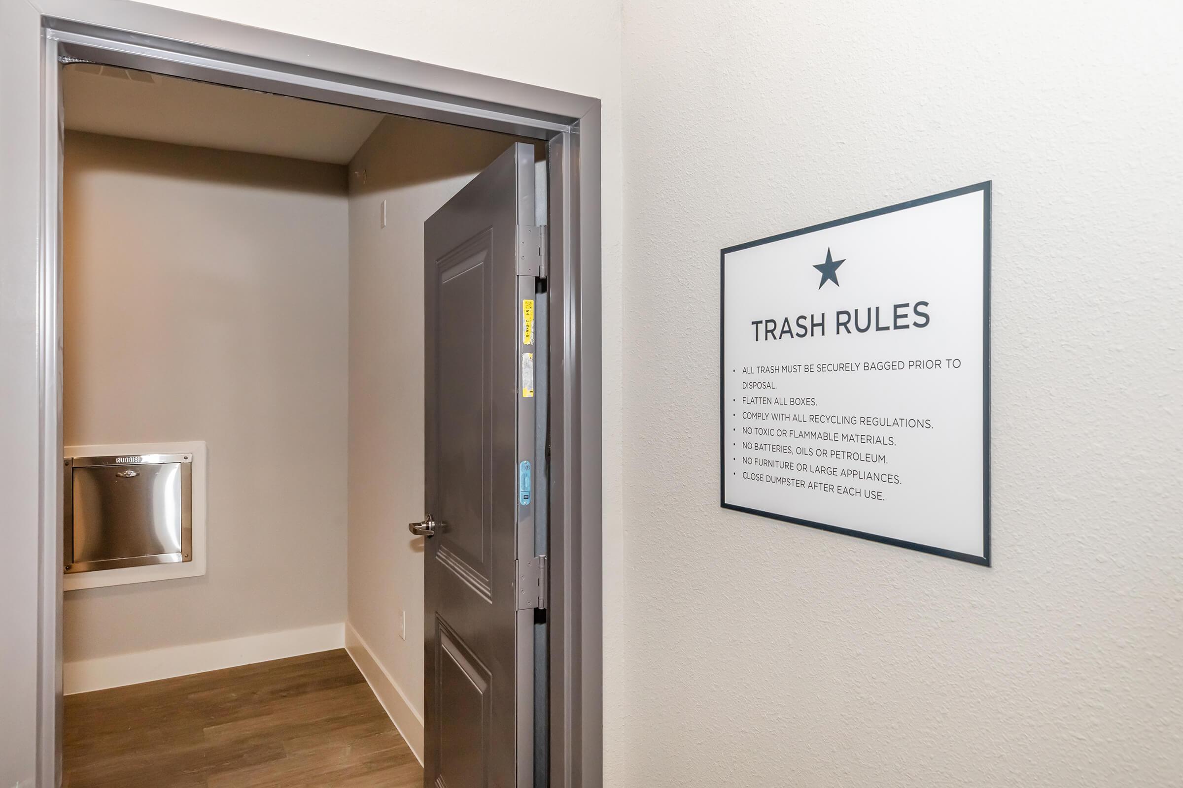 A hallway leading to a trash disposal area. A sign on the wall outlines trash rules, including guidelines for proper disposal and prohibited materials. The space is finished with light-colored walls and a clean floor, creating a modern and tidy environment. A door leads further into the area.