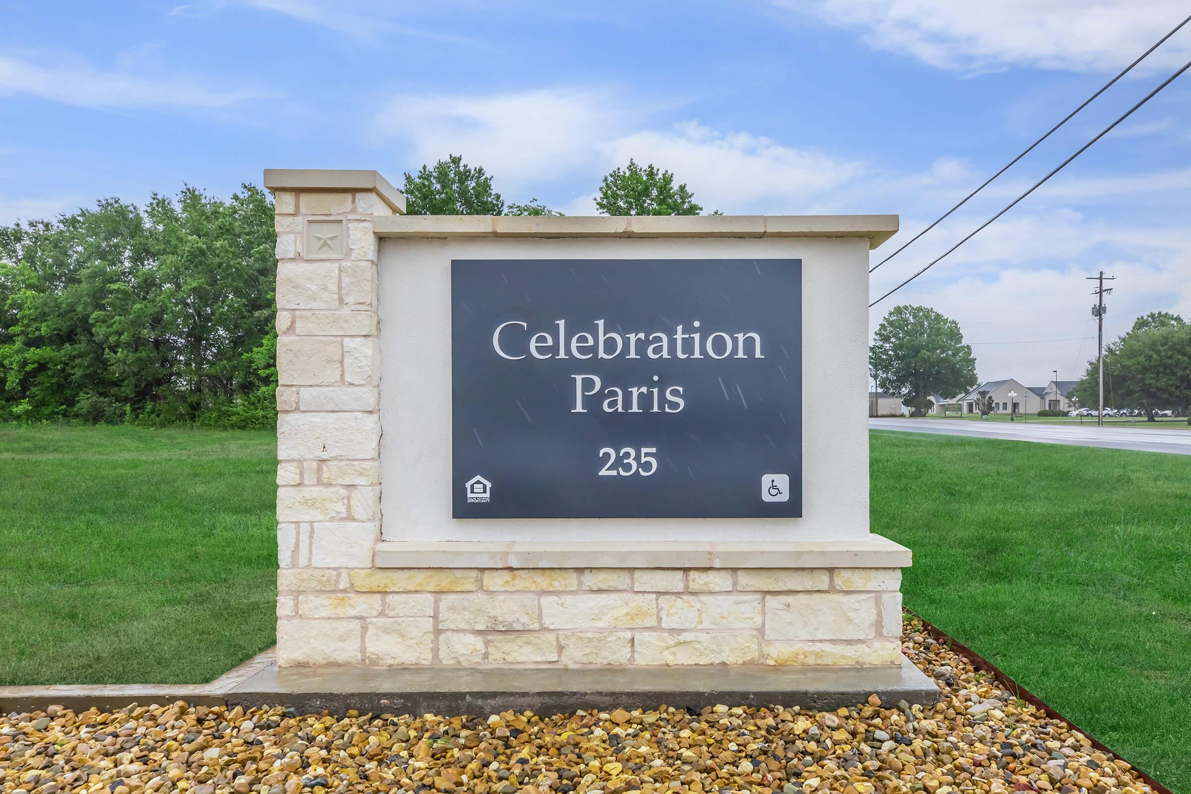 Sign displaying "Celebration Paris" with the number "235," set against a backdrop of green grass and trees. The sign features a stone base and a dark panel for the text, indicating a welcoming location.