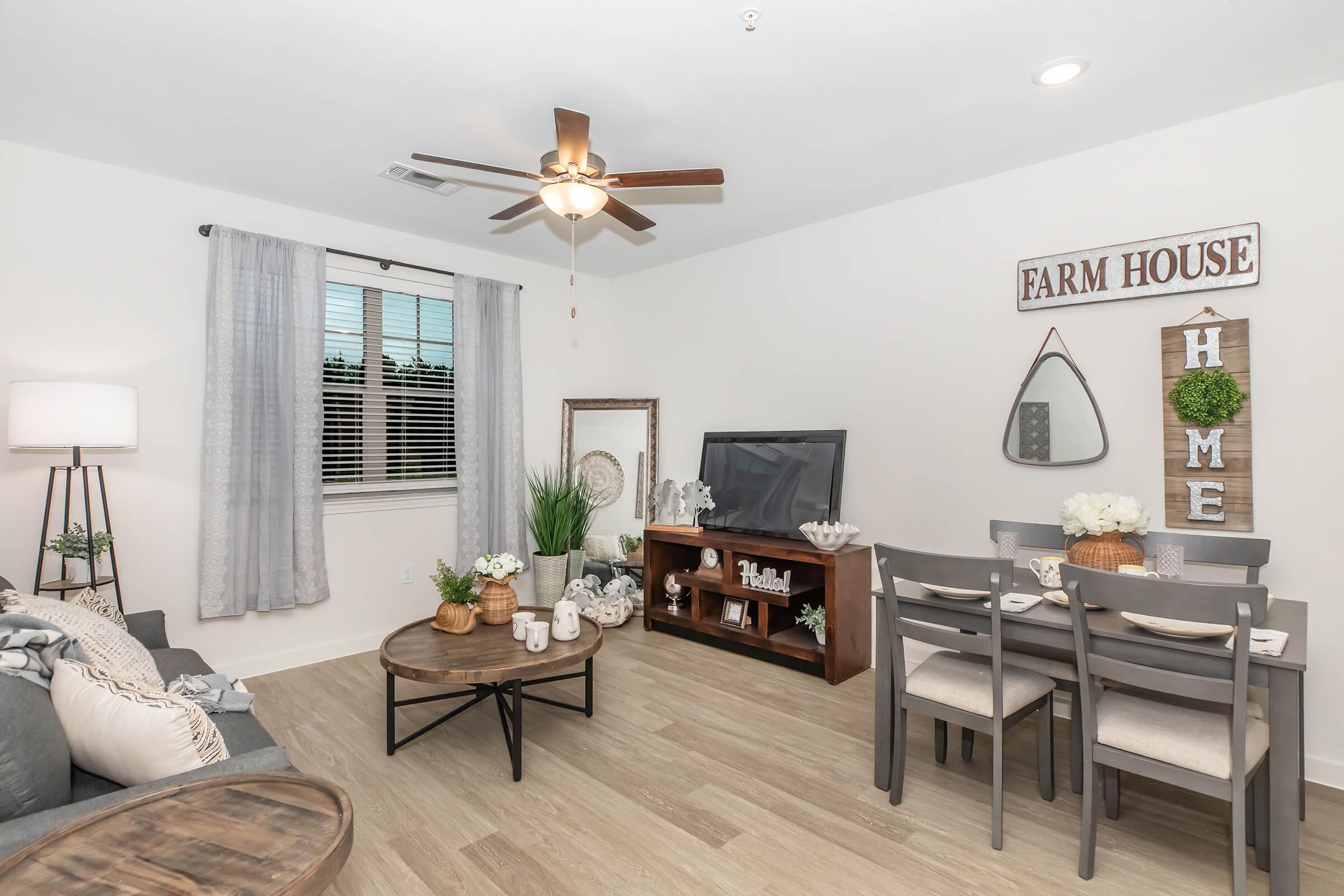 Cozy living room with a gray sofa, a round wooden coffee table, and a light wood dining table with chairs. Decor includes a potted plant, wall art with "FARM HOUSE" and "HOME," a television, and curtains. The space is bright with natural light and a ceiling fan.