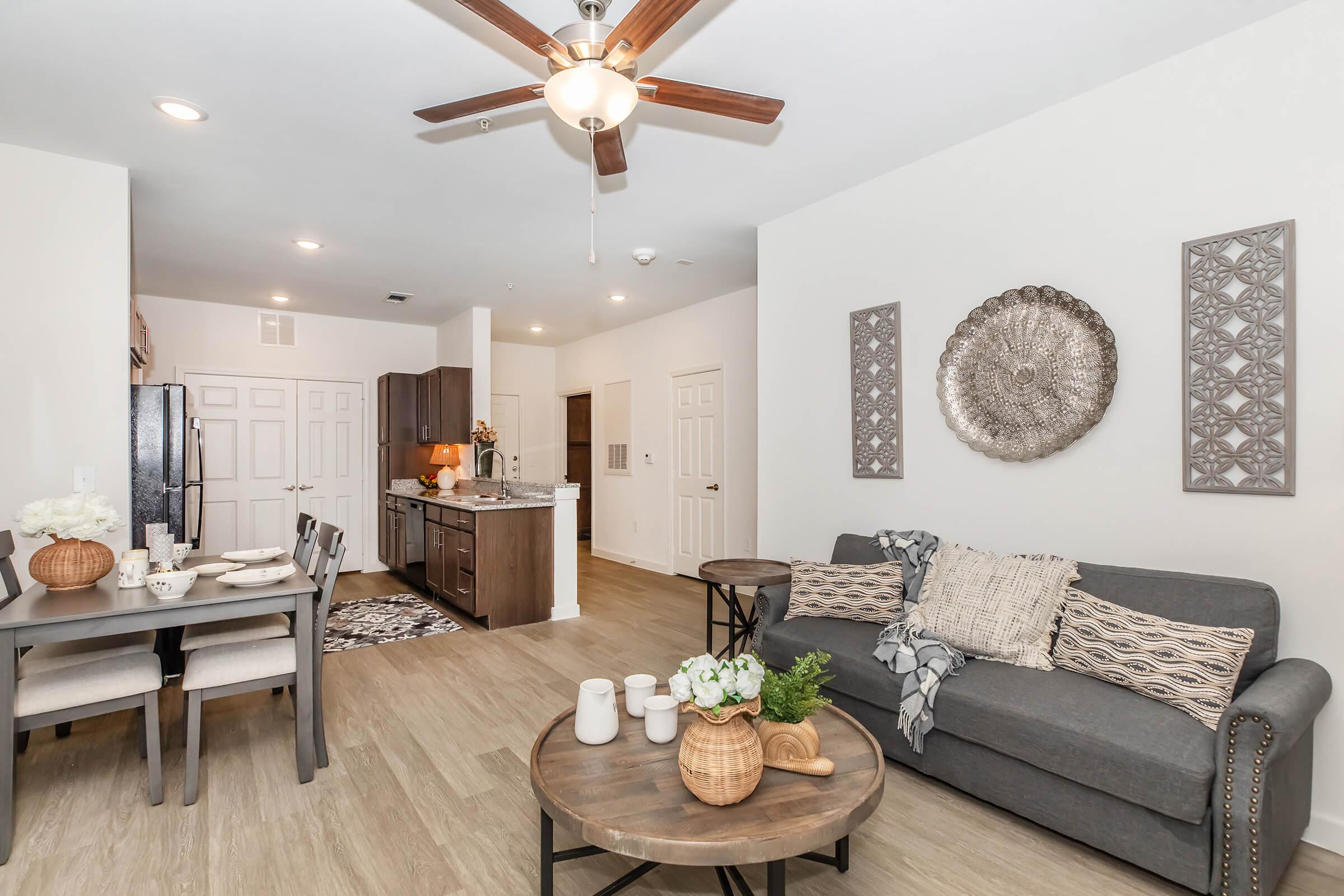 A modern living area featuring a gray sofa with decorative pillows, a round coffee table with flowers, and a dining table set for four. The room has light wood flooring, a ceiling fan, and wall art. An open kitchen area is visible in the background, with dark cabinetry and stainless steel appliances.