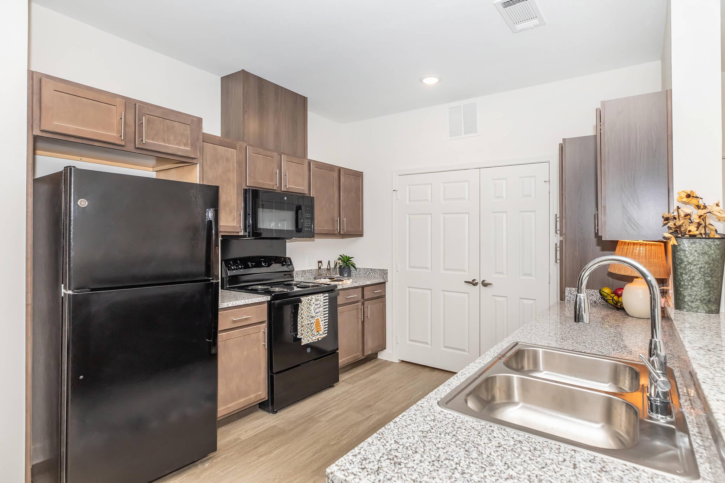 Modern kitchen featuring dark wood cabinetry, a black refrigerator, a black microwave, and a stainless steel oven. The granite countertop provides ample workspace, with a double sink and decorative items on display. Bright lighting enhances the open and inviting atmosphere.