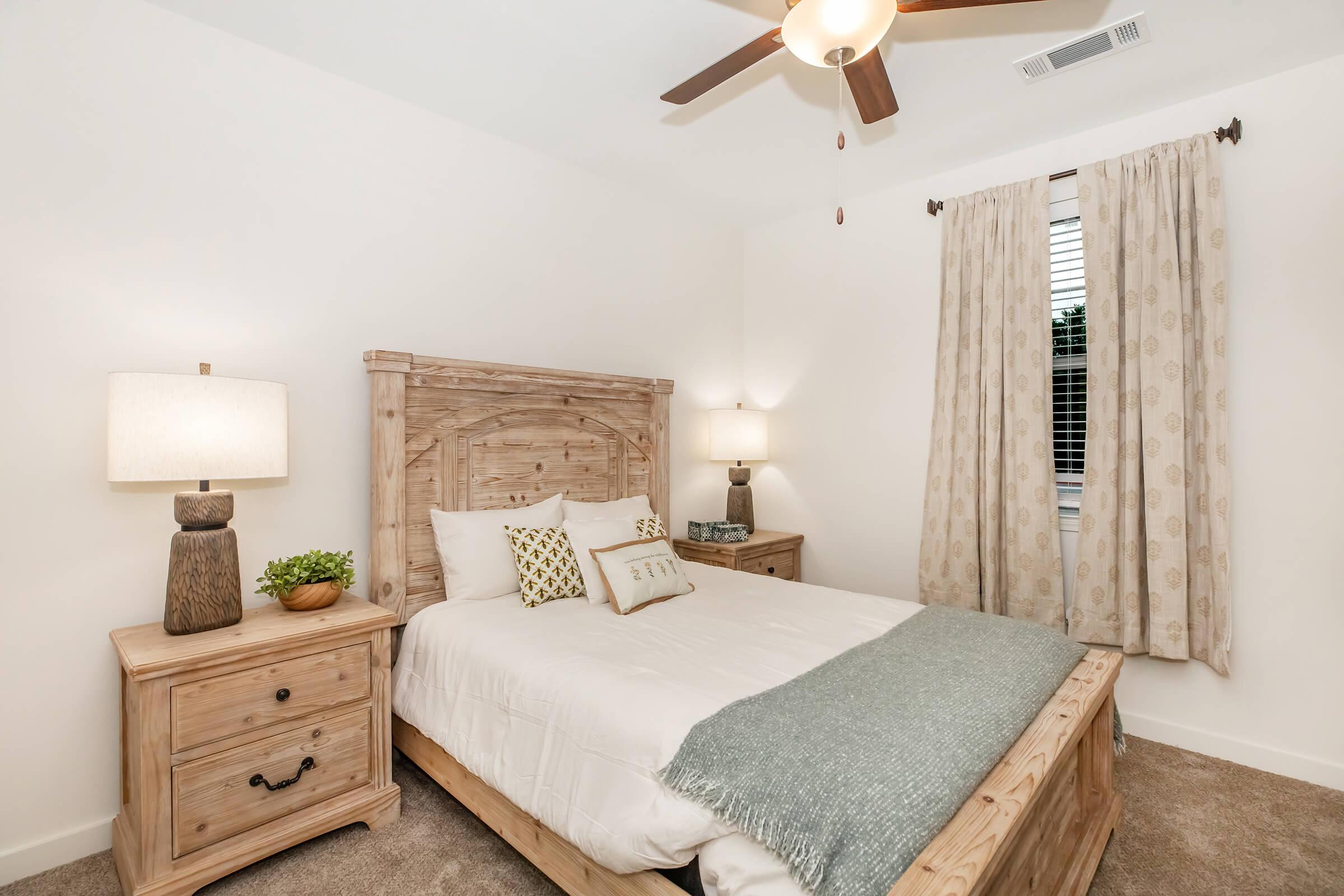 A cozy bedroom featuring a wooden bed frame with a light-colored quilt and decorative pillows. There's a nightstand on each side, one with a lamp and the other with a small plant. Soft beige curtains adorn the window, allowing natural light to brighten the space. A ceiling fan adds to the ambiance.
