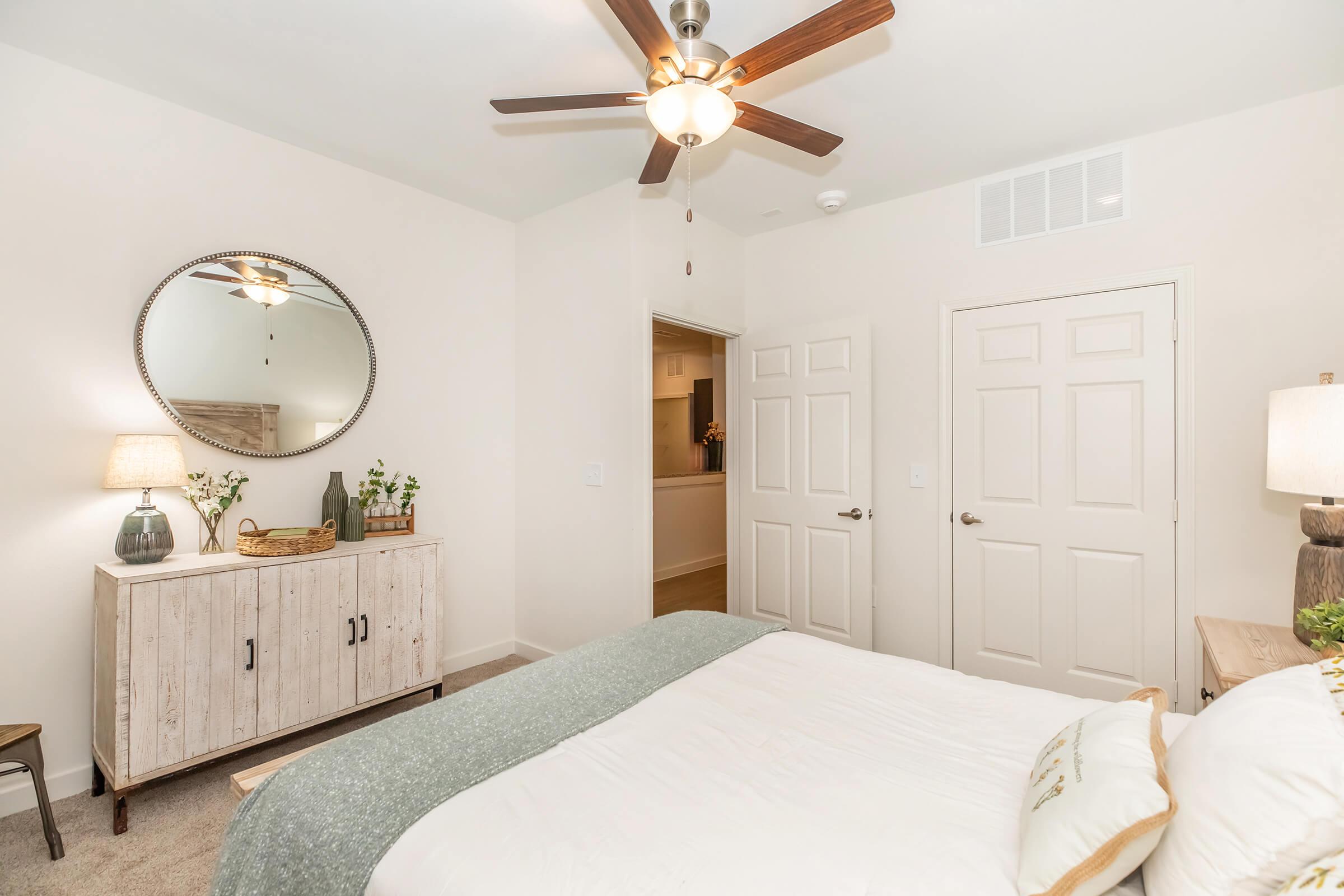 A cozy bedroom featuring a bed with white bedding and a green throw. There's a round mirror above a wooden dresser, with a lamp and decorative plants. A ceiling fan is visible, and two doors lead to other areas. The walls are painted a light color, creating a warm and inviting atmosphere.