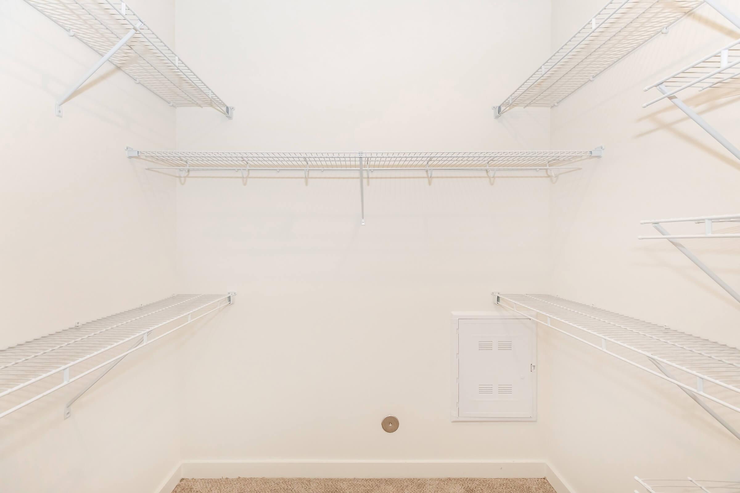 A clean, empty closet featuring white wire shelving on three sides, with a single panel on the wall for electrical access, and beige carpet flooring. The walls are plain and painted in a light color, creating a minimalistic storage space.