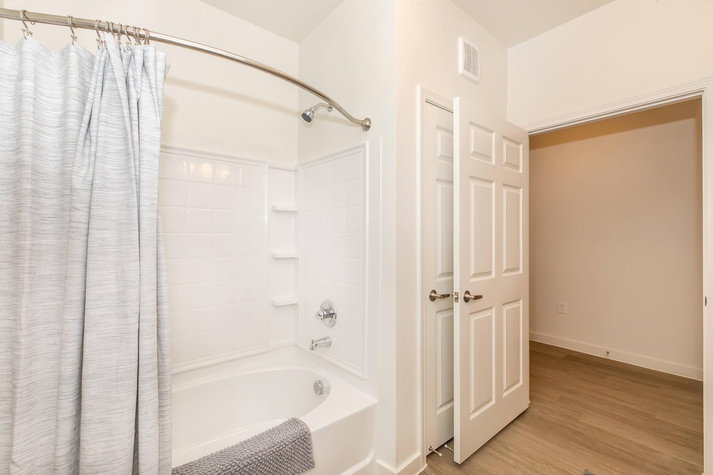 A clean bathroom featuring a bathtub with a shower curtain, a white tiled shower area, and a chrome faucet. There are built-in shelves for toiletries. To the side, a doorway leads to another room, and the floor is covered with light-colored wood laminate.