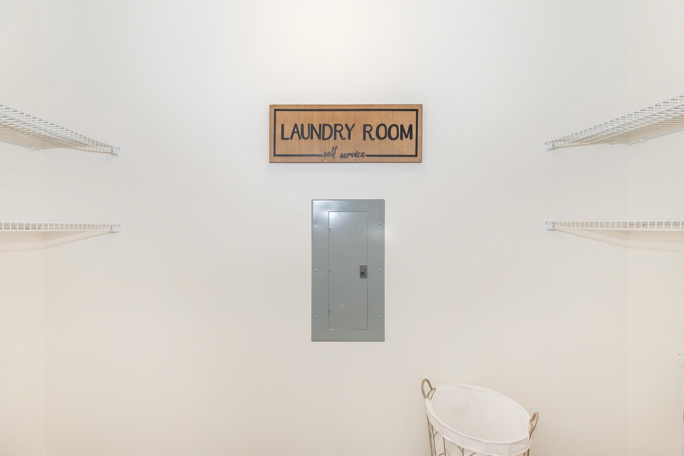 A minimalistic laundry room with a sign labeled "LAUNDRY ROOM" on the wall. To the sides, there are two wire shelves for storage. Below the sign, there's an electrical panel. A white laundry basket is positioned on the floor, creating a clean and organized space.