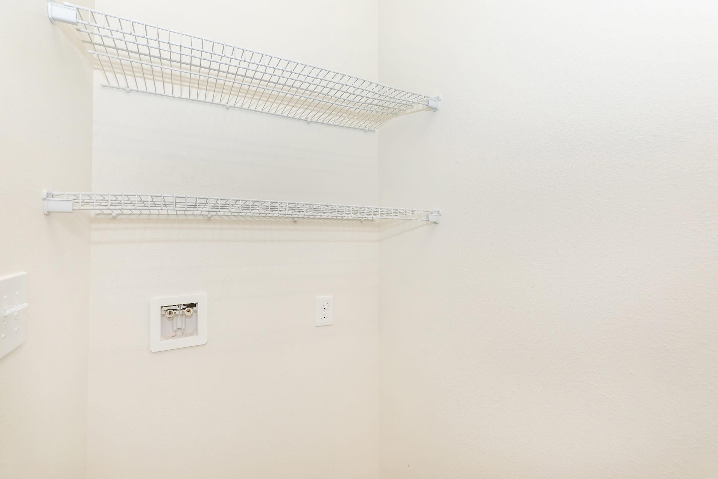 Empty room with two white wire shelves mounted on a light beige wall. There is an electrical outlet and a small wall mount, but the space is otherwise unoccupied, creating a clean and minimalistic appearance.