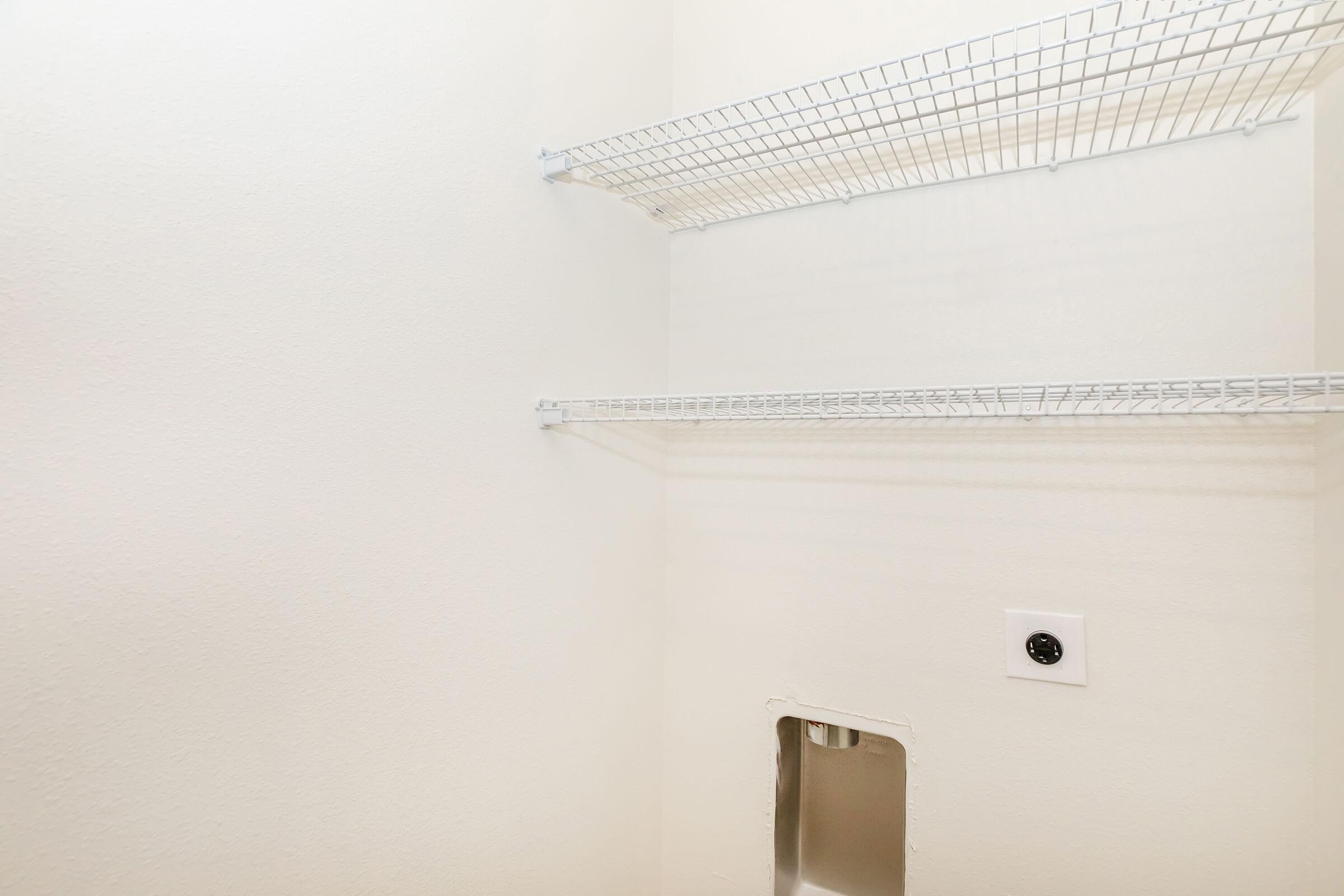 An empty closet with two wire shelving units mounted on the wall. The walls are plain and white, creating a minimalist appearance. There is an electrical outlet visible near the floor, and a small opening or recess in the wall. The overall space looks neat and uncluttered.