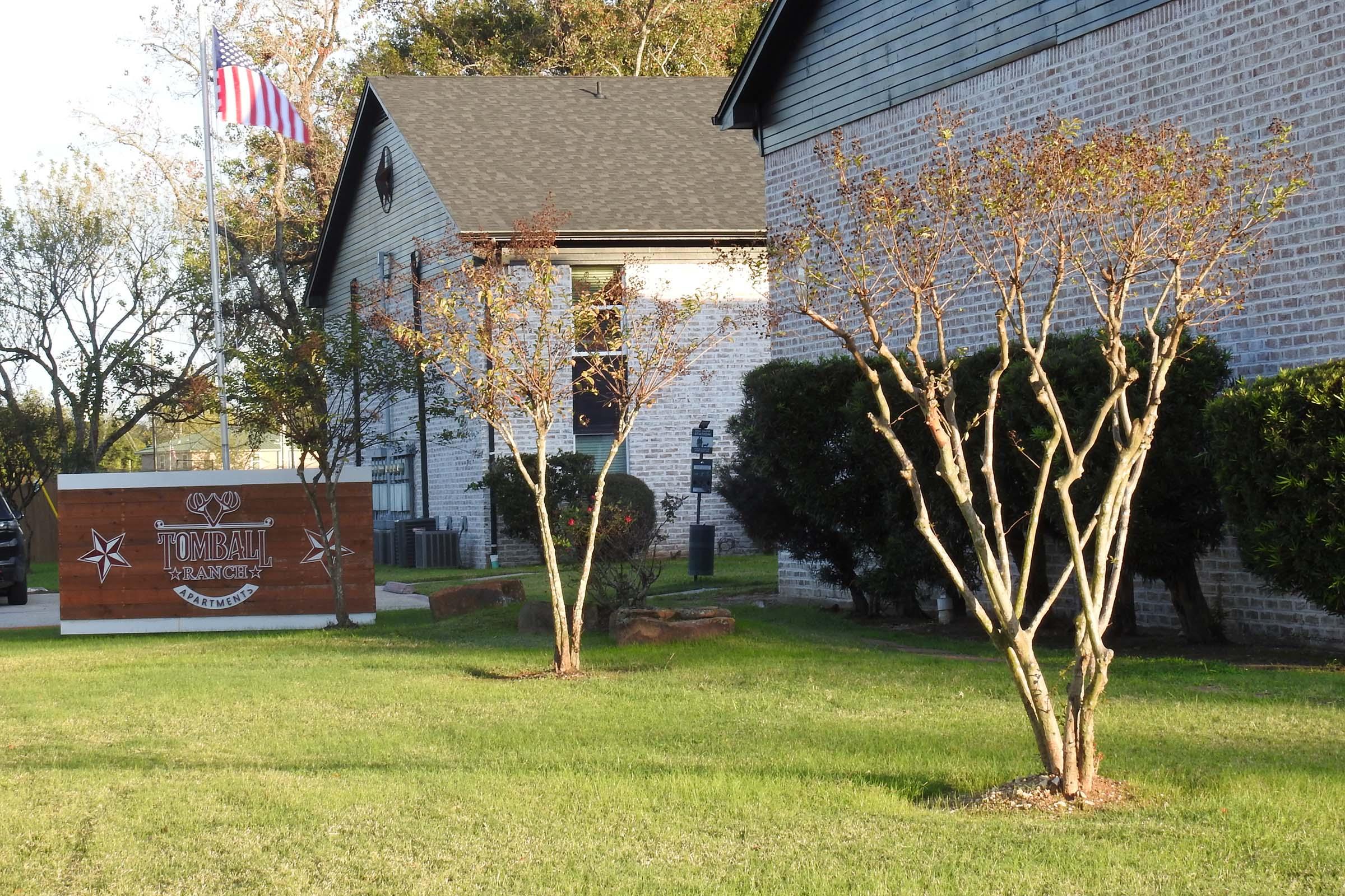 A building with a brick exterior and a sign that reads "Tomball Trailers" is visible along with a small American flag. In the foreground, there are two young trees and a well-maintained green lawn, creating a tidy landscape. The scene is well-lit, suggesting it is late afternoon or early evening.