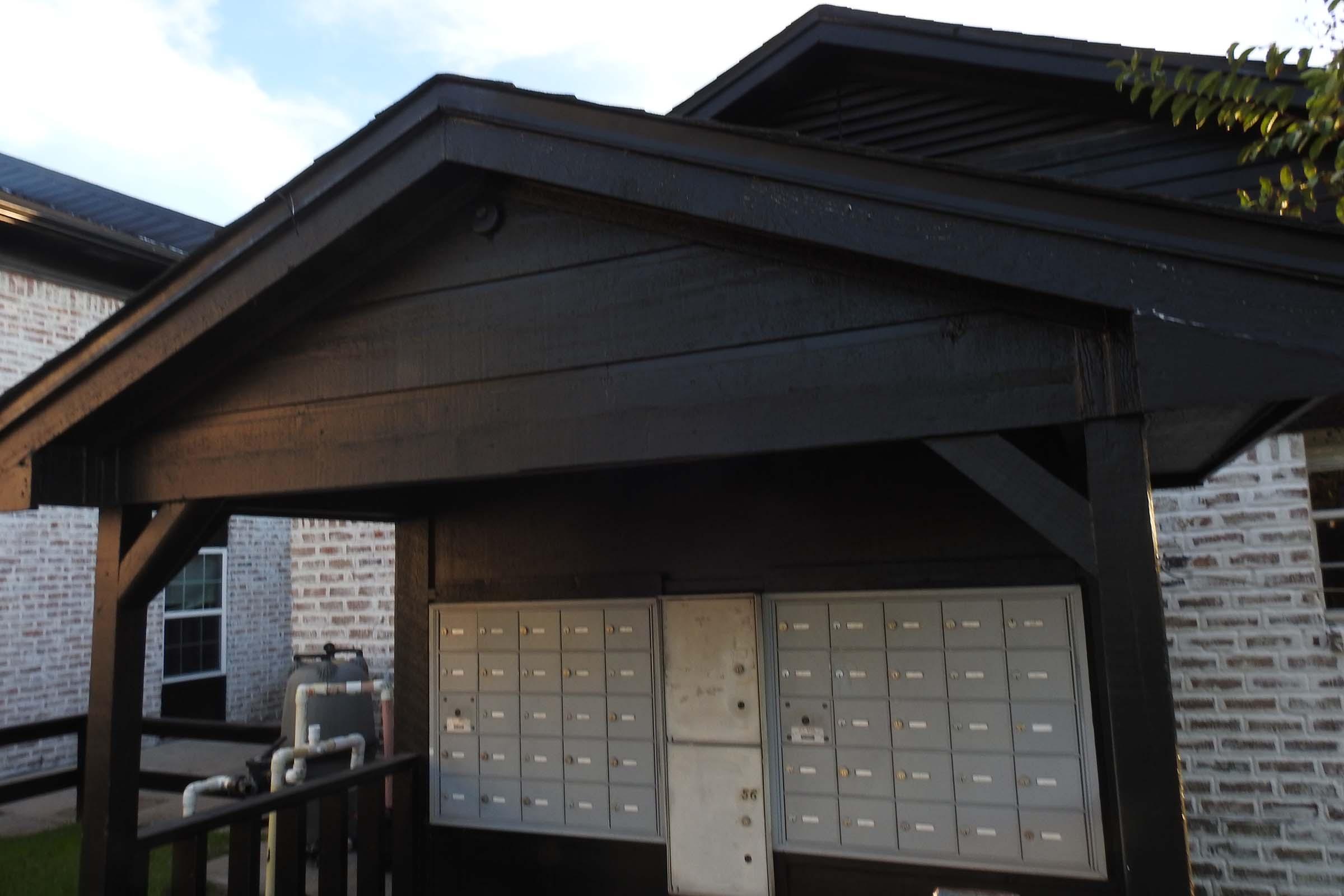 Post office box area with a black wooden structure and multiple mailbox compartments. The roof extends over the mailboxes, and there are visible utility features nearby. The building is adjacent to a brick wall with a light blue sky in the background.