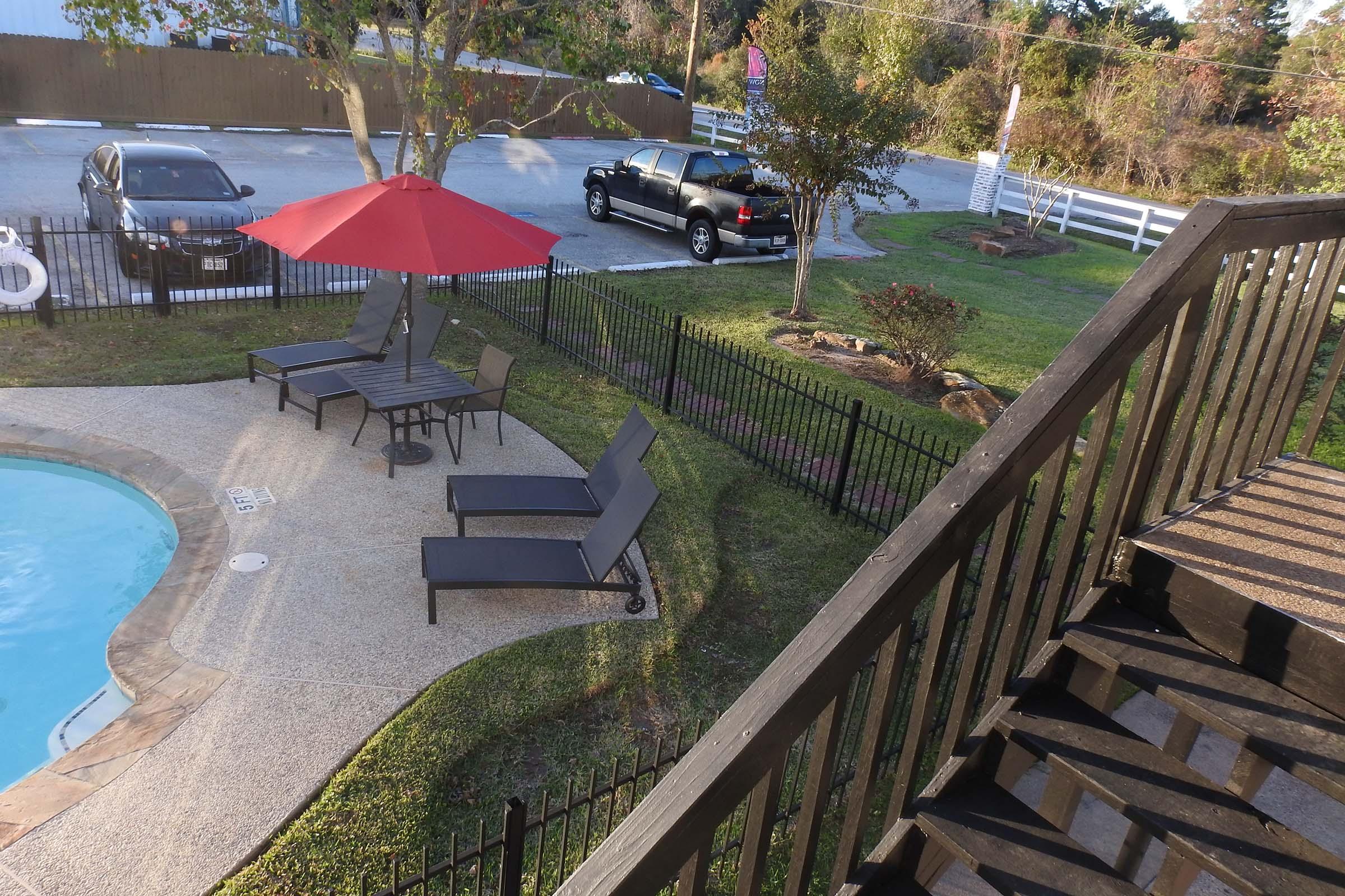 A view from a balcony overlooking a pool area with loungers and a table under a red umbrella. In the background, parked cars are visible, along with trees and a grassy area. The scene is set in a residential or recreational environment.