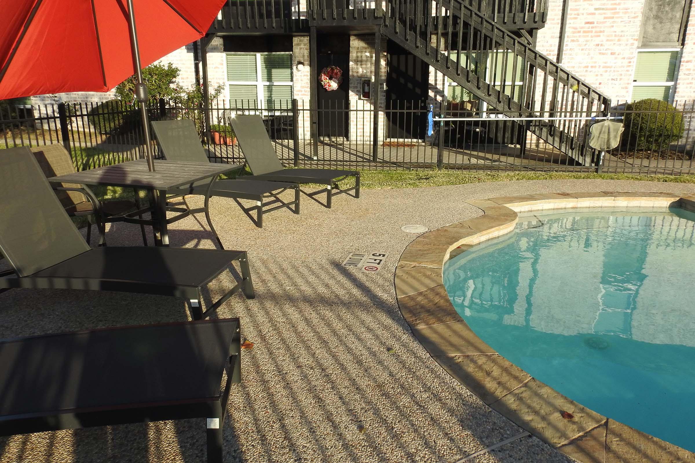 A sunny poolside scene featuring a clear blue swimming pool with a shallow end marked at 5 feet. There are several black lounge chairs and a table under a bright red umbrella. In the background, a multi-story building with a staircase and a wreath on the door is visible, along with a fenced area.